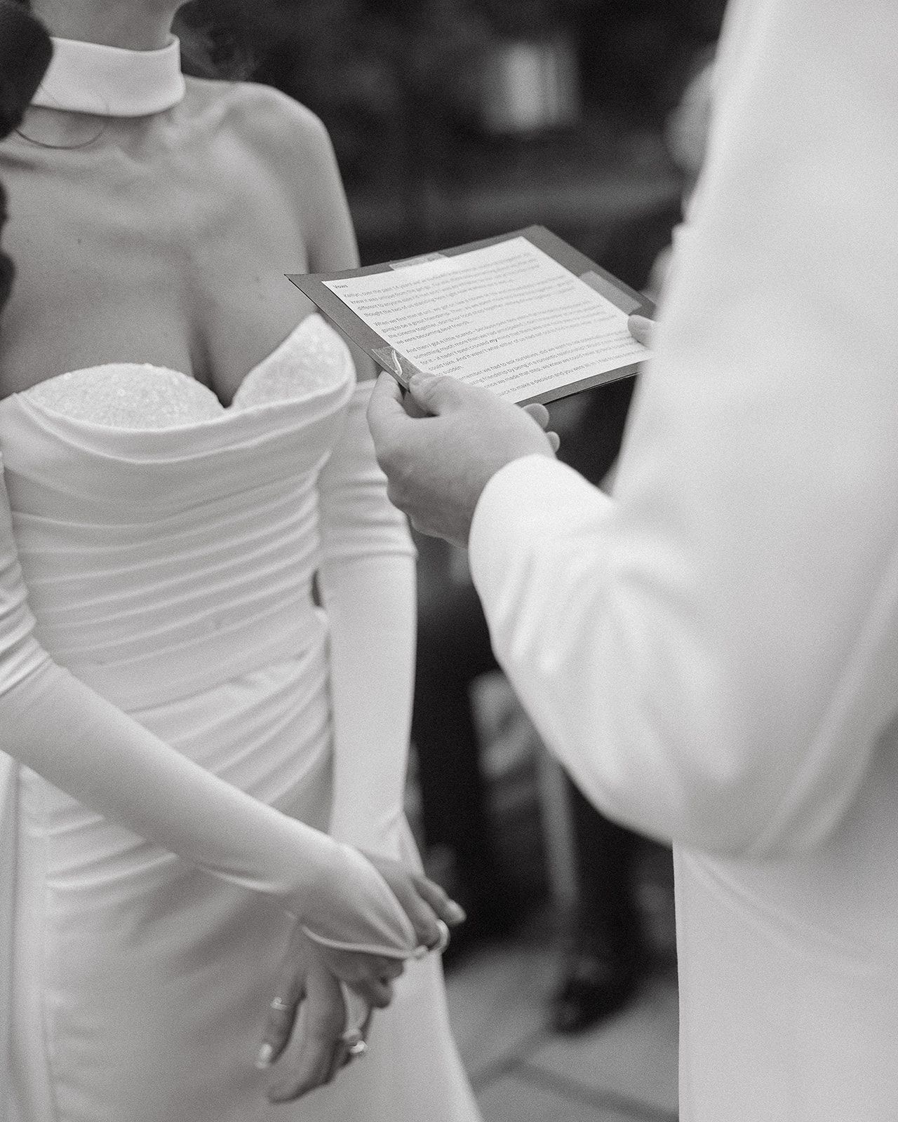A close-up black and white shot of a couple during their wedding ceremony as vows are read from a paper.
