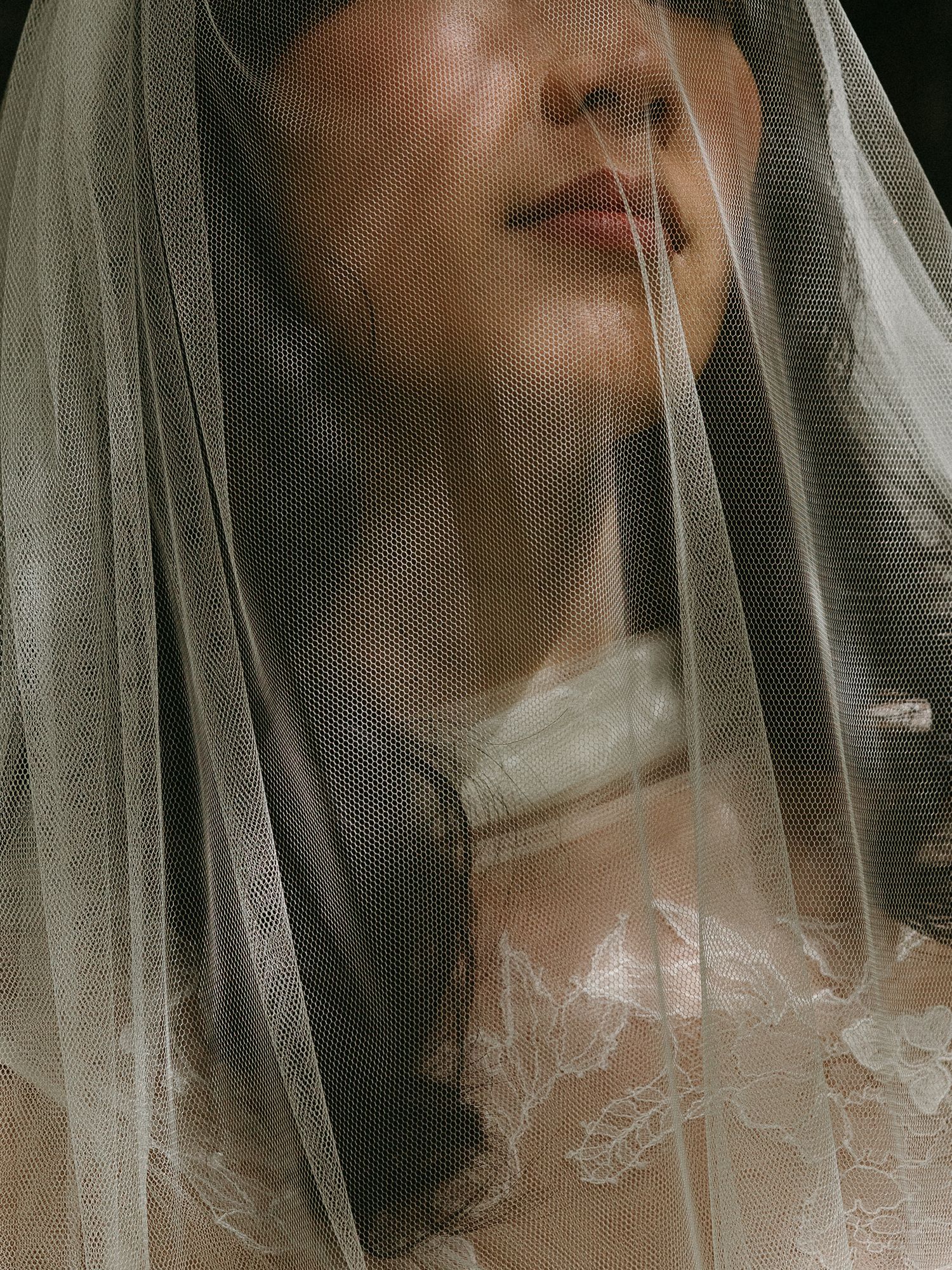 A bride wearing a delicate lace wedding dress, her face partially veiled by sheer tulle fabric.