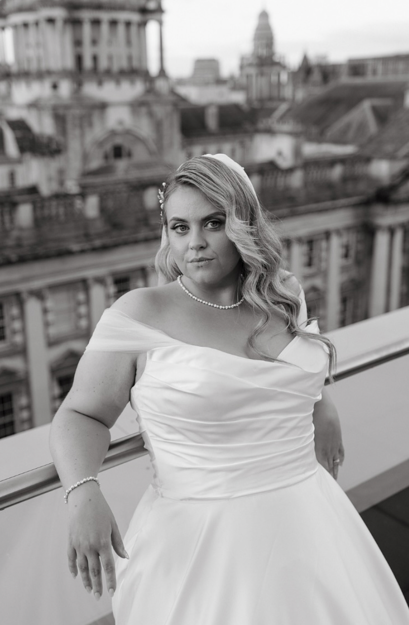 A black and white bridal portrait showcasing an off-shoulder wedding gown against an architectural city backdrop.