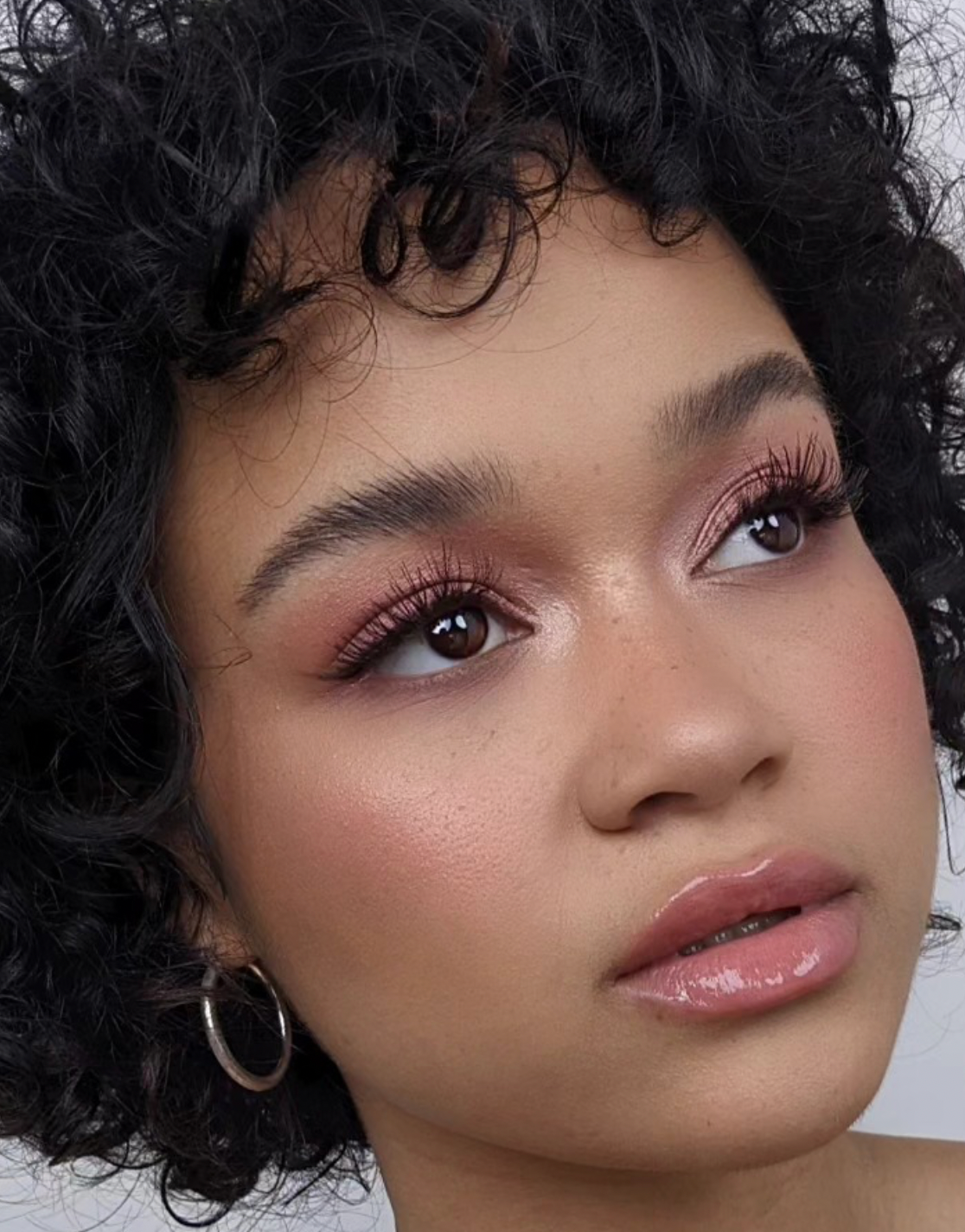 A beauty portrait showcasing pink eyeshadow, glossy lips and natural curly hair against a white background.