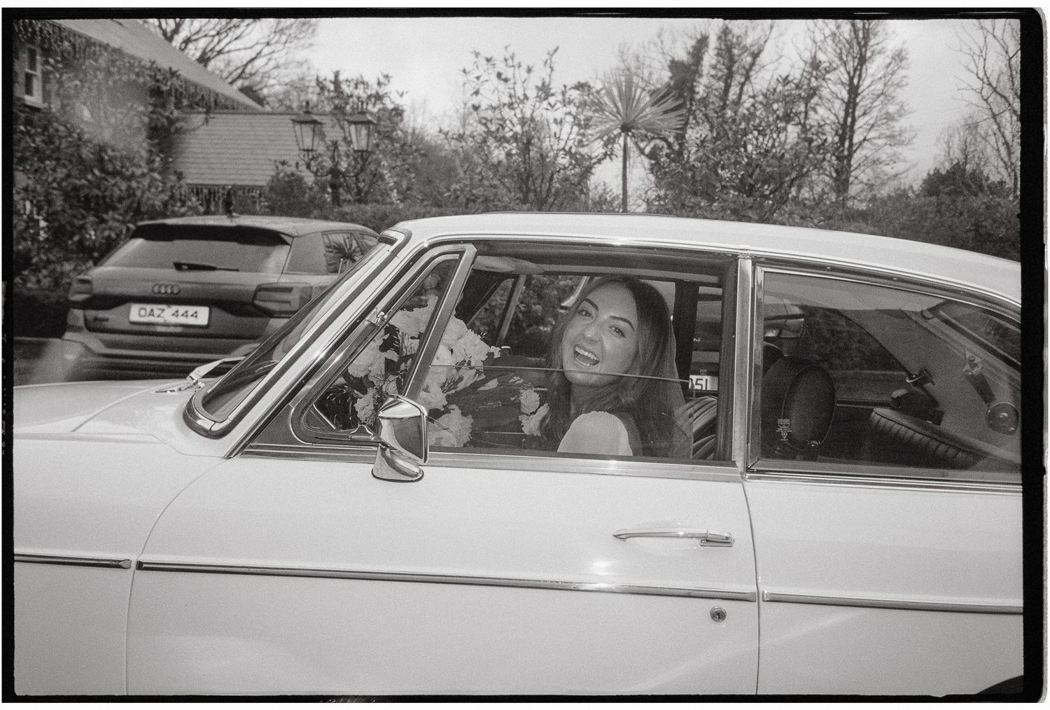 Black and white photograph showing a person sitting in the driver's seat of a vintage car, viewed through the side window.