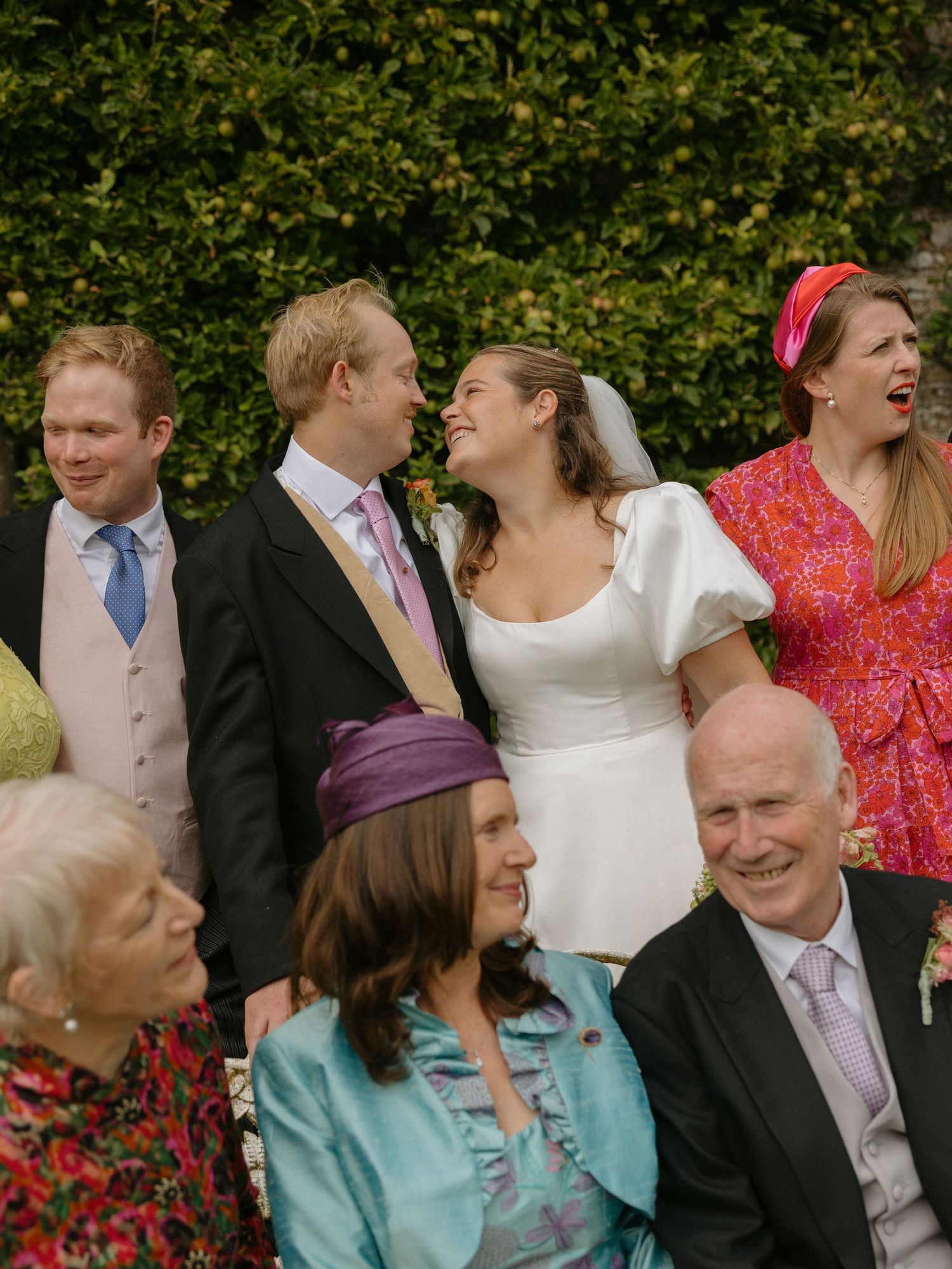 A group of wedding guests share joyful laughter together in front of lush green foliage.