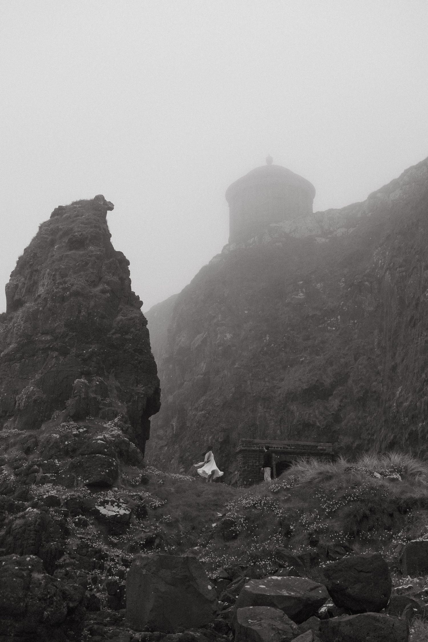 Black and white photo of a couple in wedding attire on rocky cliffs near a misty stone tower in foggy weather.