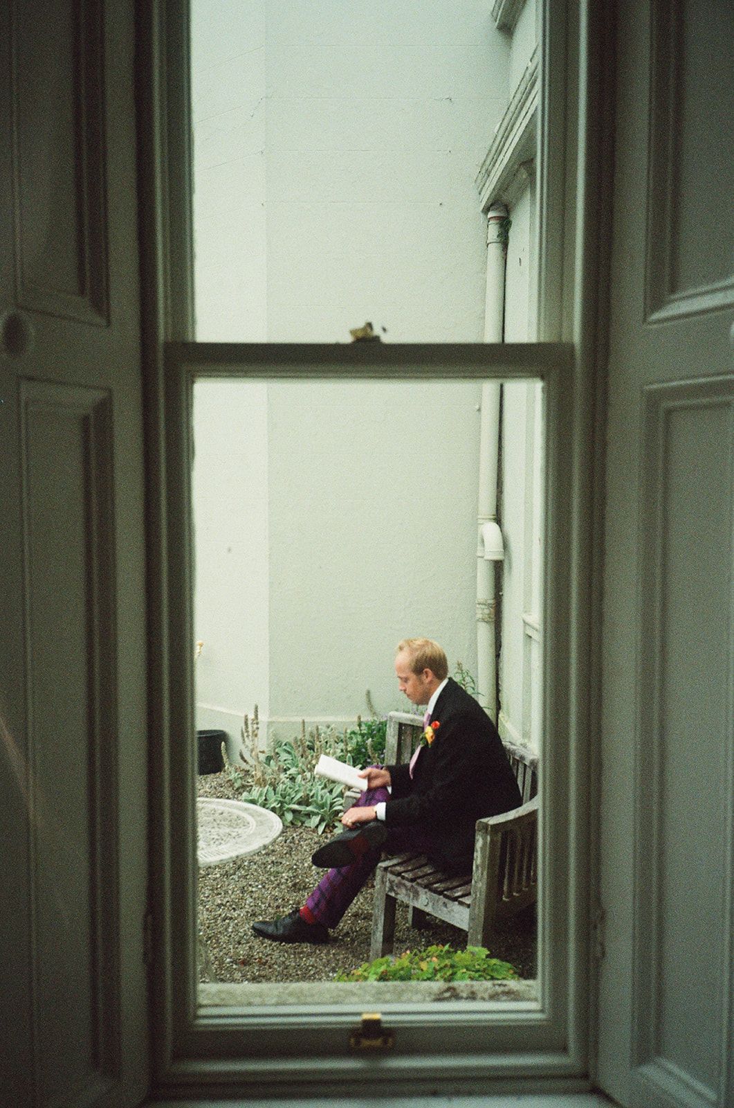 Through a window view, a person in dark clothing sits alone on a bench outside a light-colored building.
