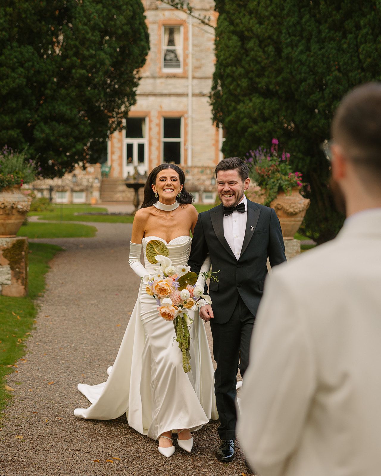 A couple walking down a garden path at their wedding ceremony in front of a historic stone building.