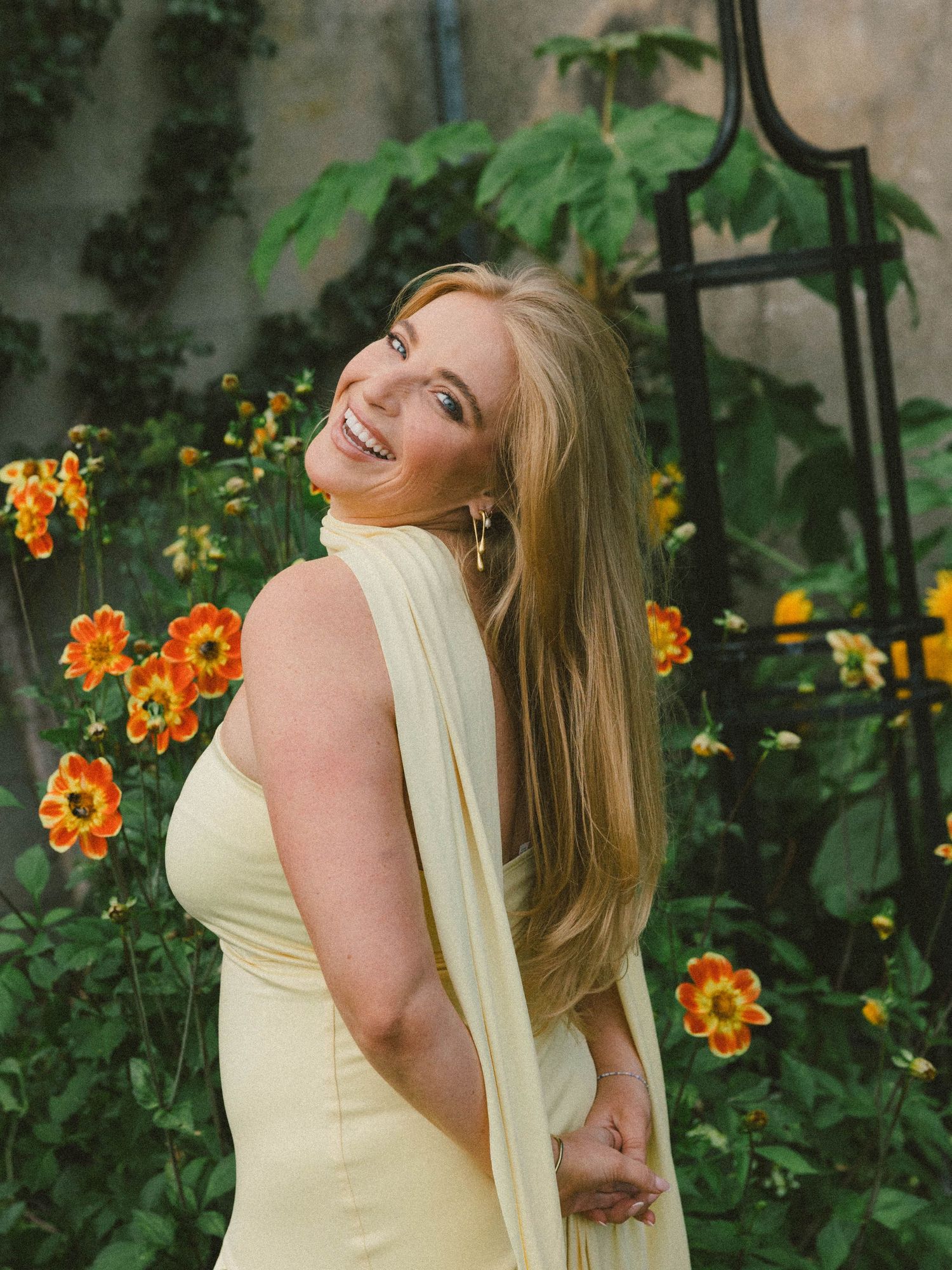A woman in a pale yellow dress poses joyfully against a garden backdrop with orange flowers.