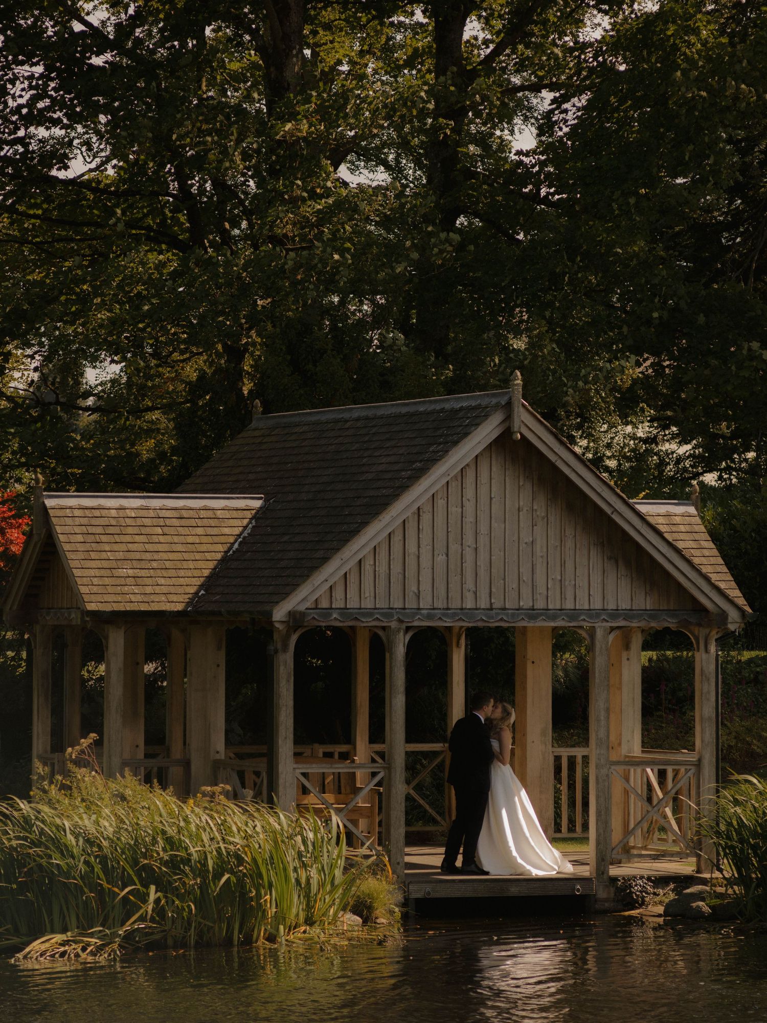 Wedding couple stands in a rustic lakeside gazebo surrounded by trees at dusk.
