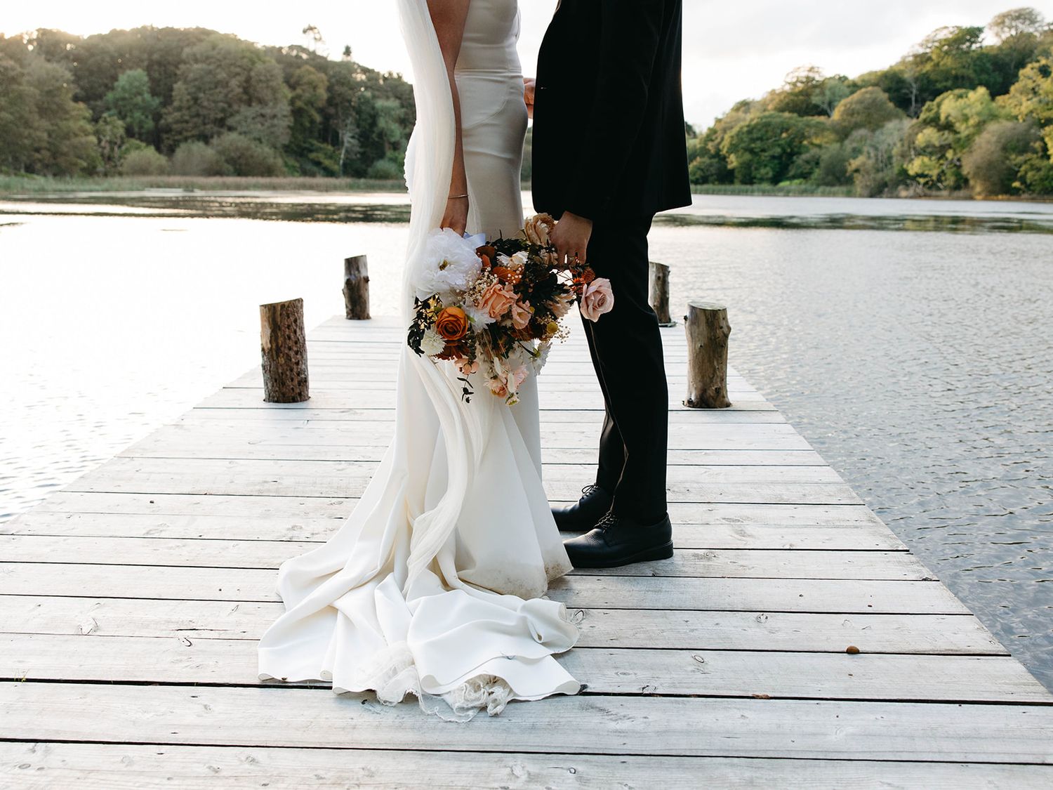 A couple stands together on a wooden dock by a lake, with the bride holding an autumn-colored bouquet.