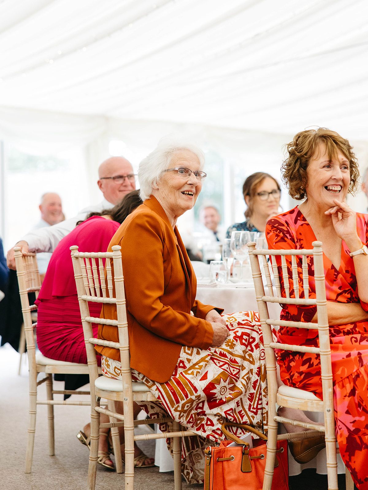 Wedding guests in colorful attire laugh and smile while seated in elegant white chairs at a reception celebration.