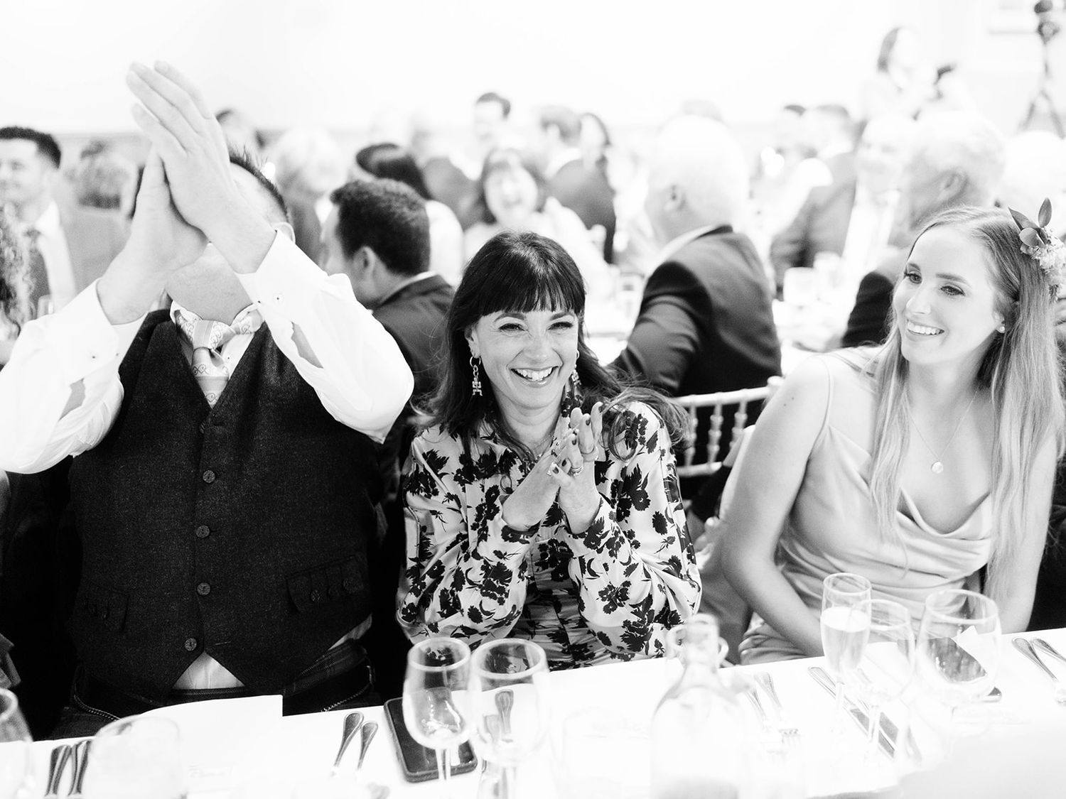 Wedding guests share laughter at a reception table as one guest covers his face in a candid black and white photo.