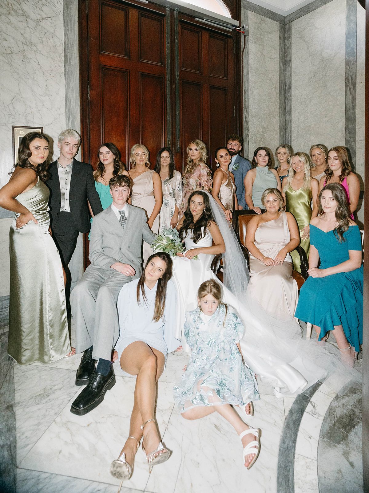 Wedding party posing on marble stairs in formal attire including blue, white and teal dresses.