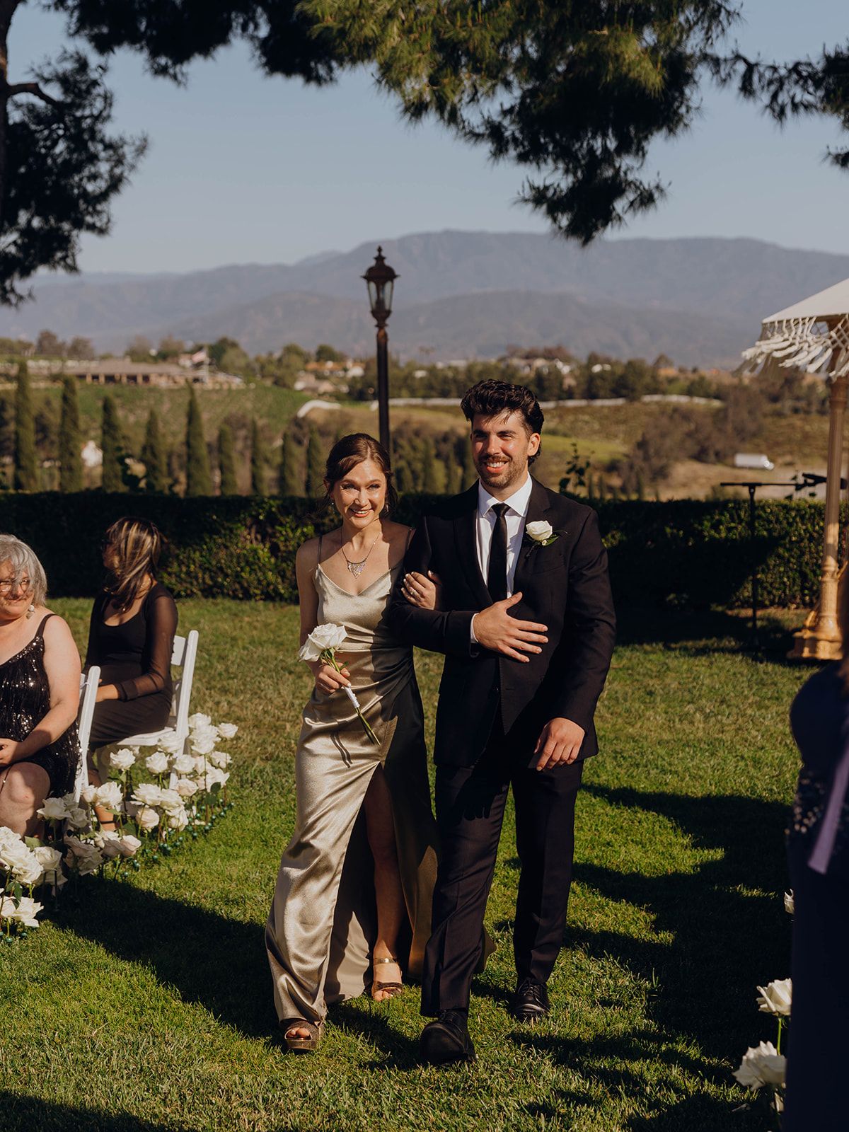 A wedding ceremony takes place on a scenic lawn with mountains in the background on a sunny afternoon.