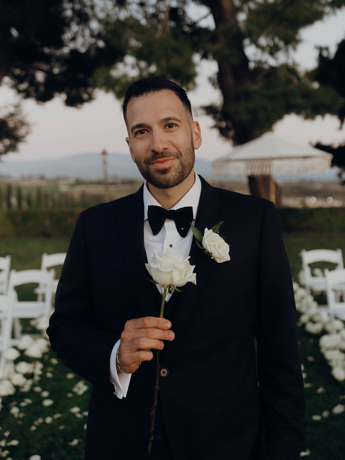 A groom in a black tuxedo and bow tie holds a white rose boutonniere at an outdoor wedding venue with chairs.