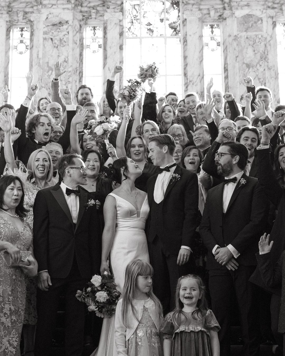 A large group of wedding guests in formal attire laugh and celebrate together in a black and white photograph.