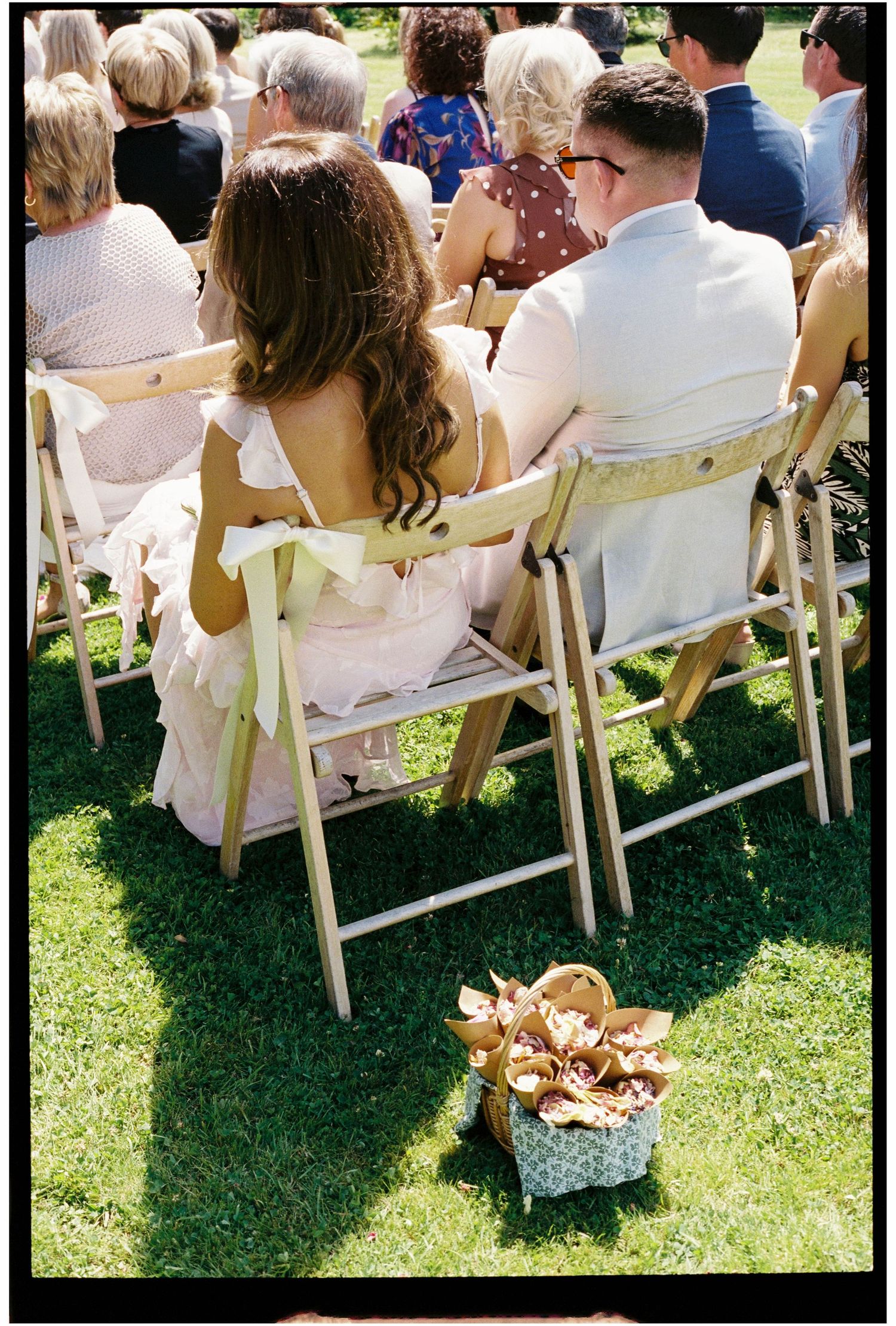 Wedding guests seated on wooden folding chairs on green grass with flower petal basket nearby.