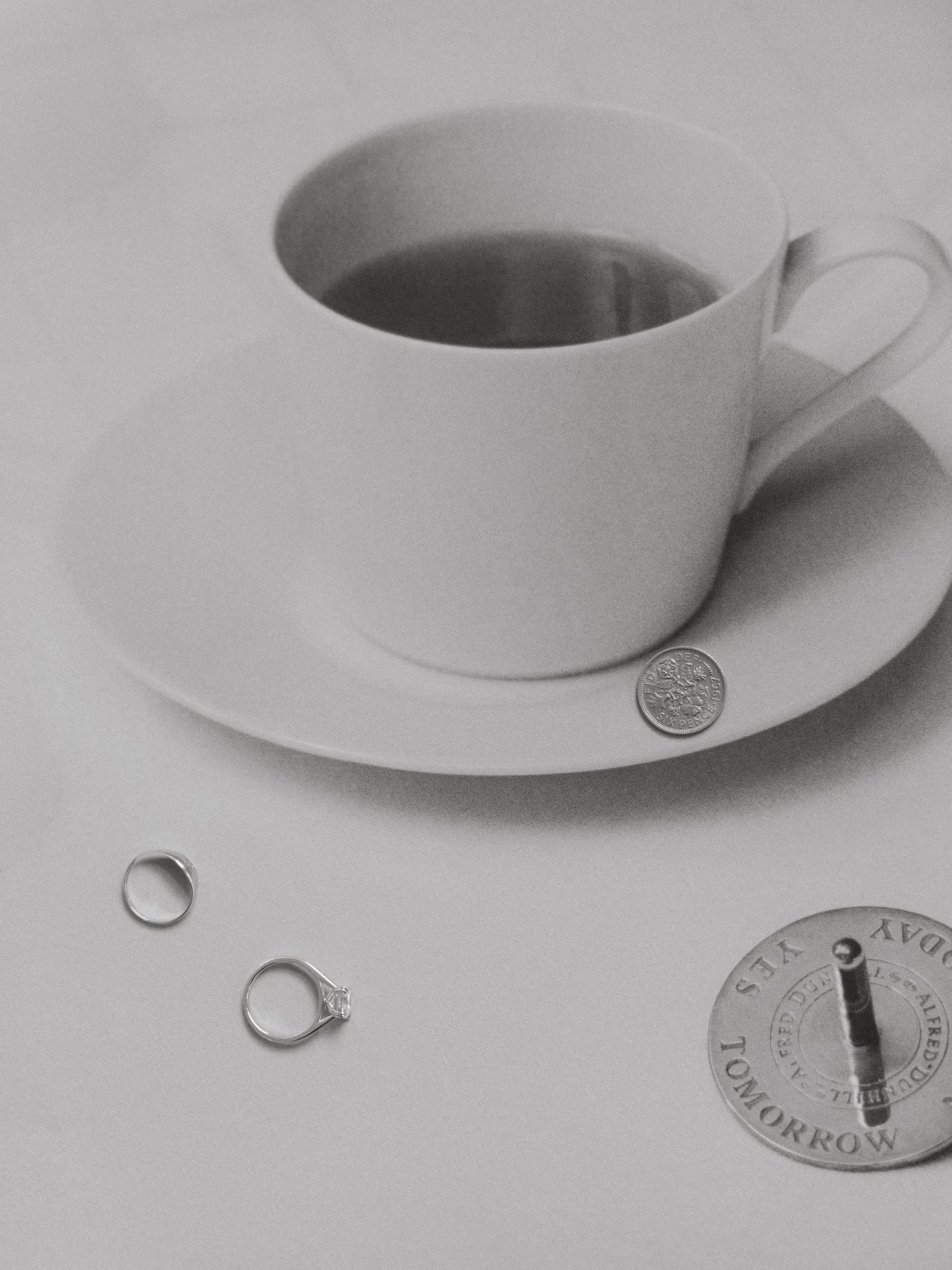 Black and white photo of a coffee cup on saucer with two rings, a coin, and a stamp beside it.