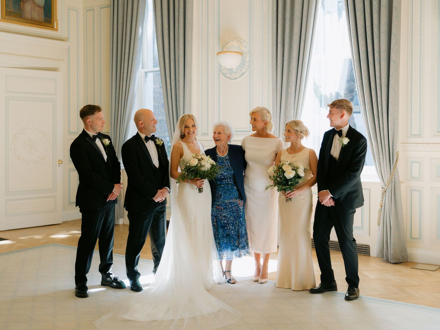 Wedding party lined up for formal portrait photos in an elegant room with blue curtains and white walls.