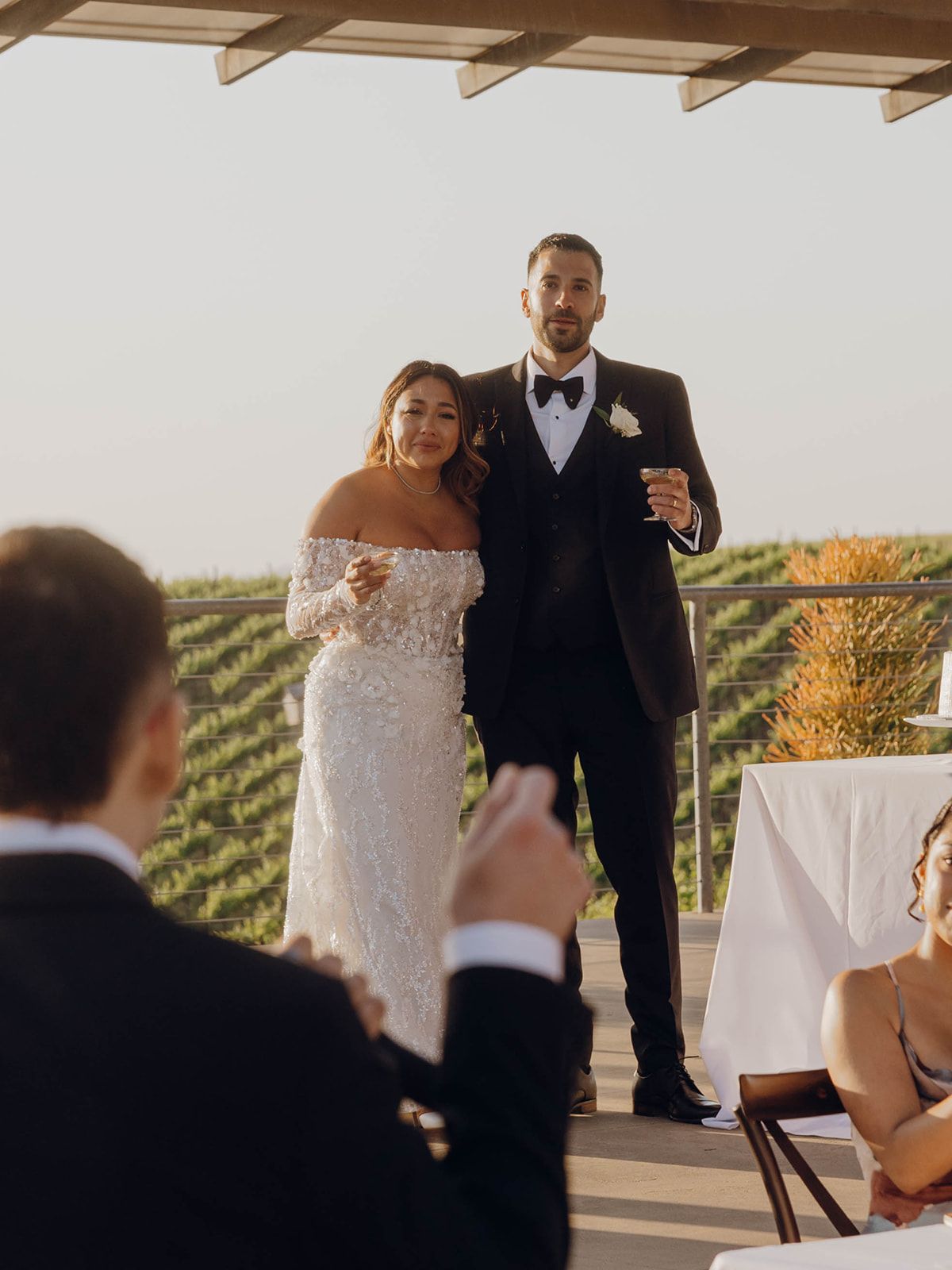 A wedding couple shares a toast at their outdoor winery ceremony with views of rolling vineyard hills in the background.