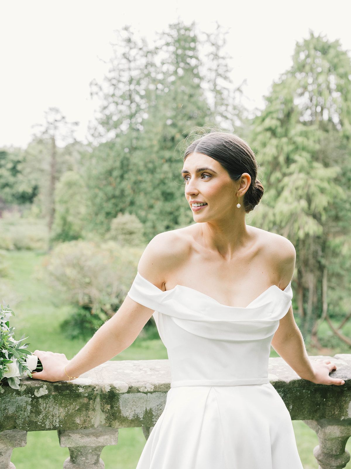 A bride in an elegant off-shoulder white wedding dress stands at a stone wall outdoors with lush greenery in the background.