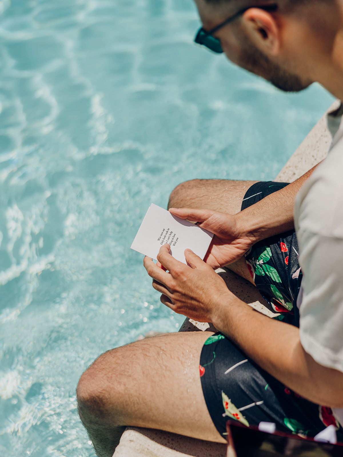 Person in swim shorts reading a letter while sitting by a blue swimming pool.