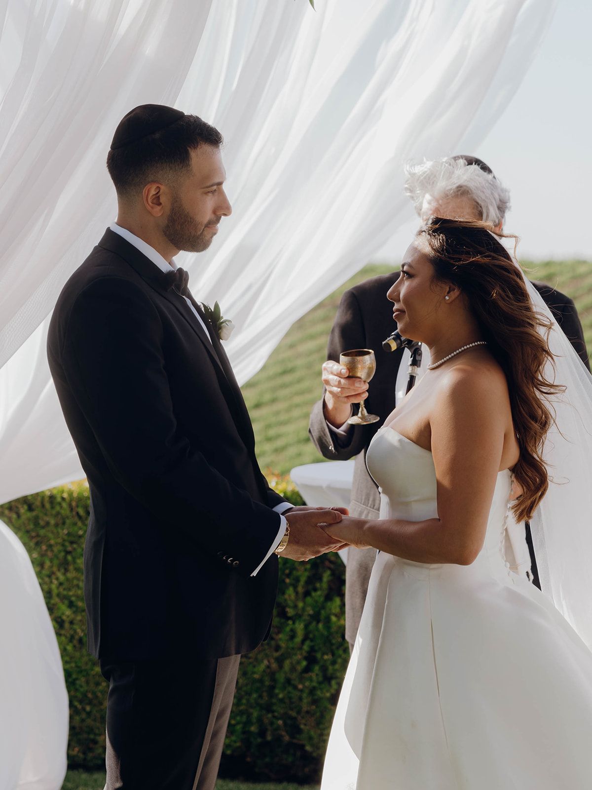 A couple exchanges wedding vows in an elegant outdoor ceremony framed by flowing white curtains and manicured hedges.