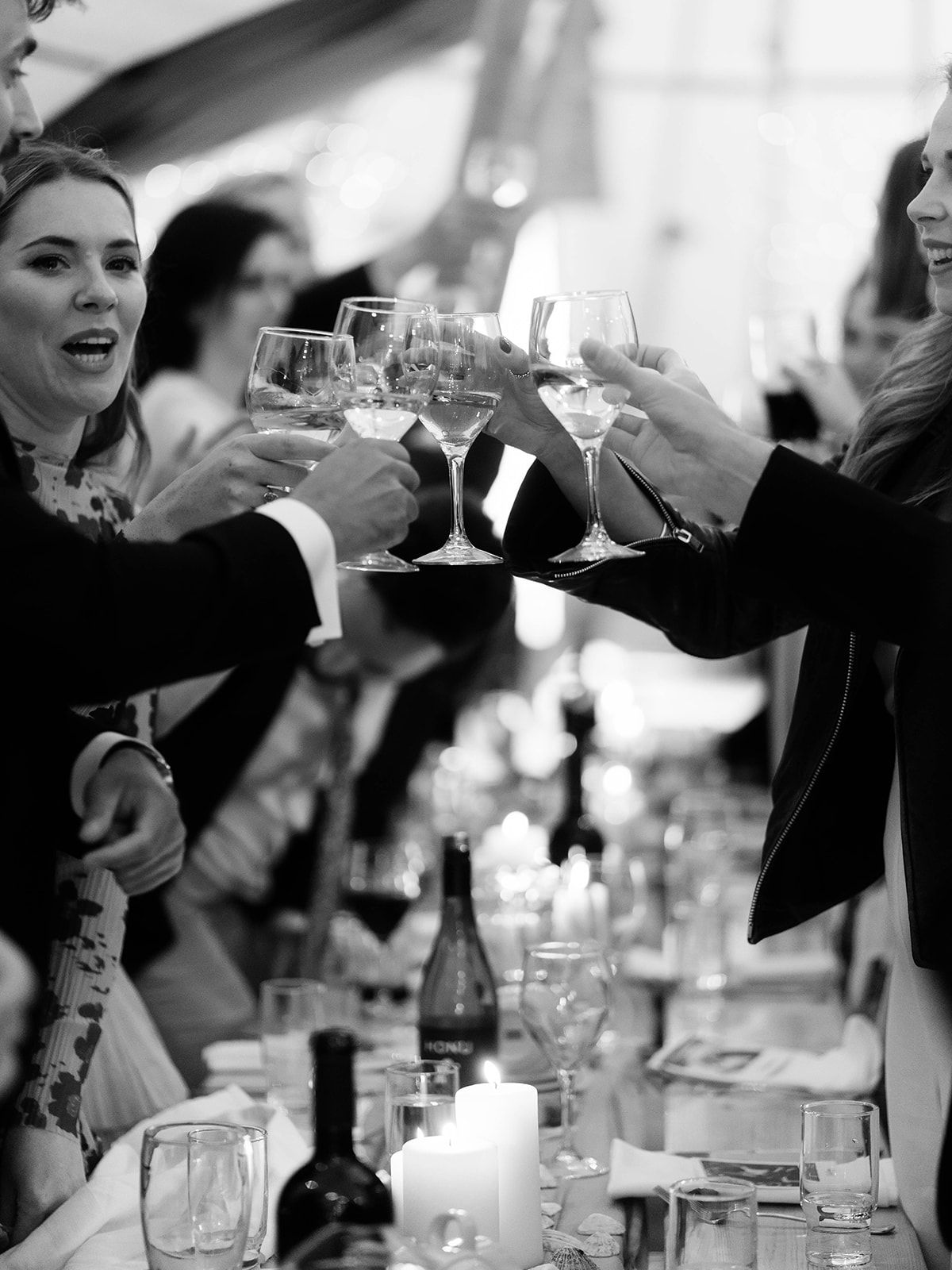Black and white photo of wedding guests toasting with wine glasses at candlelit reception table.
