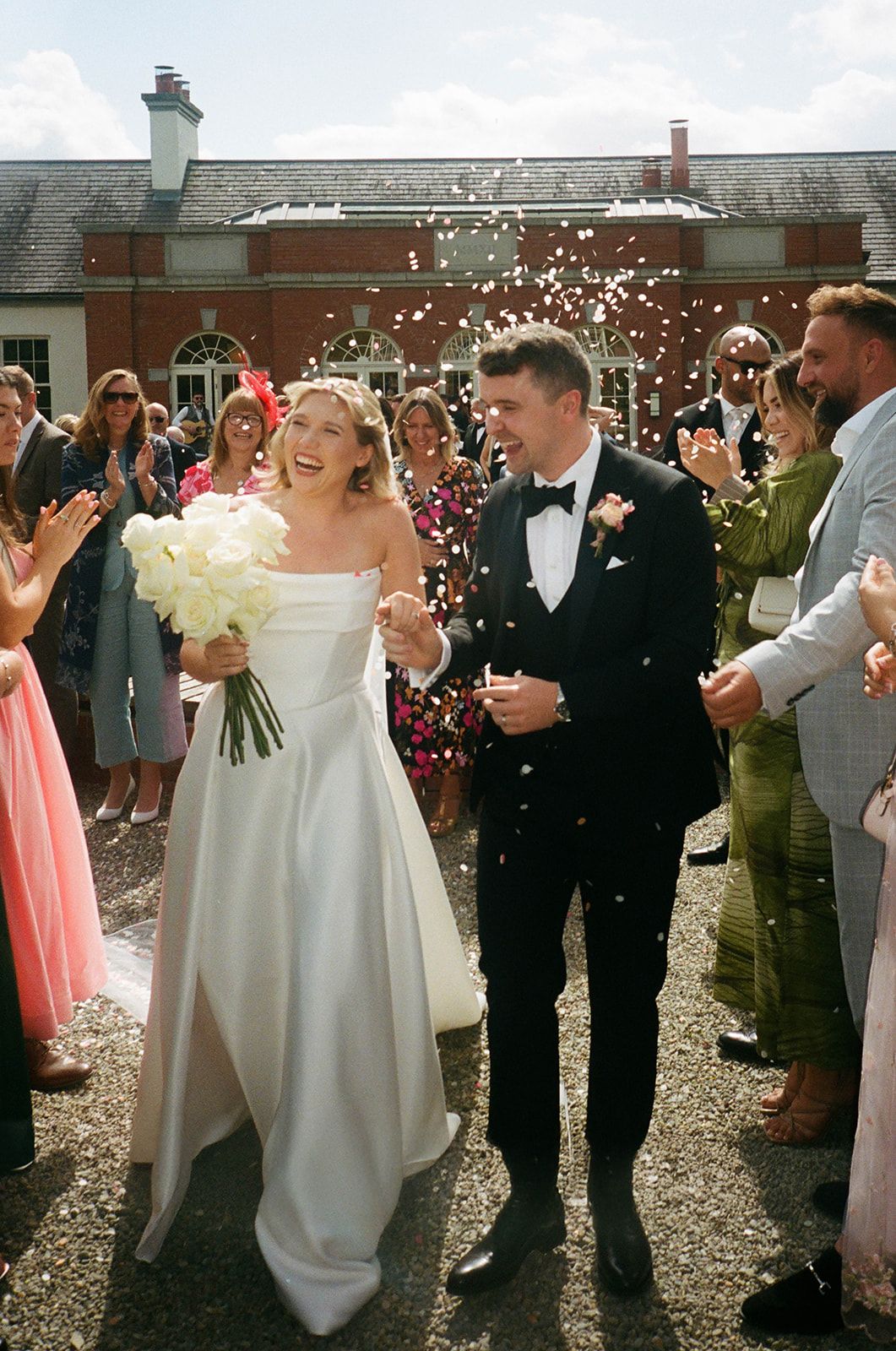 A joyful wedding celebration with confetti being thrown at a couple exiting a venue with guests cheering.