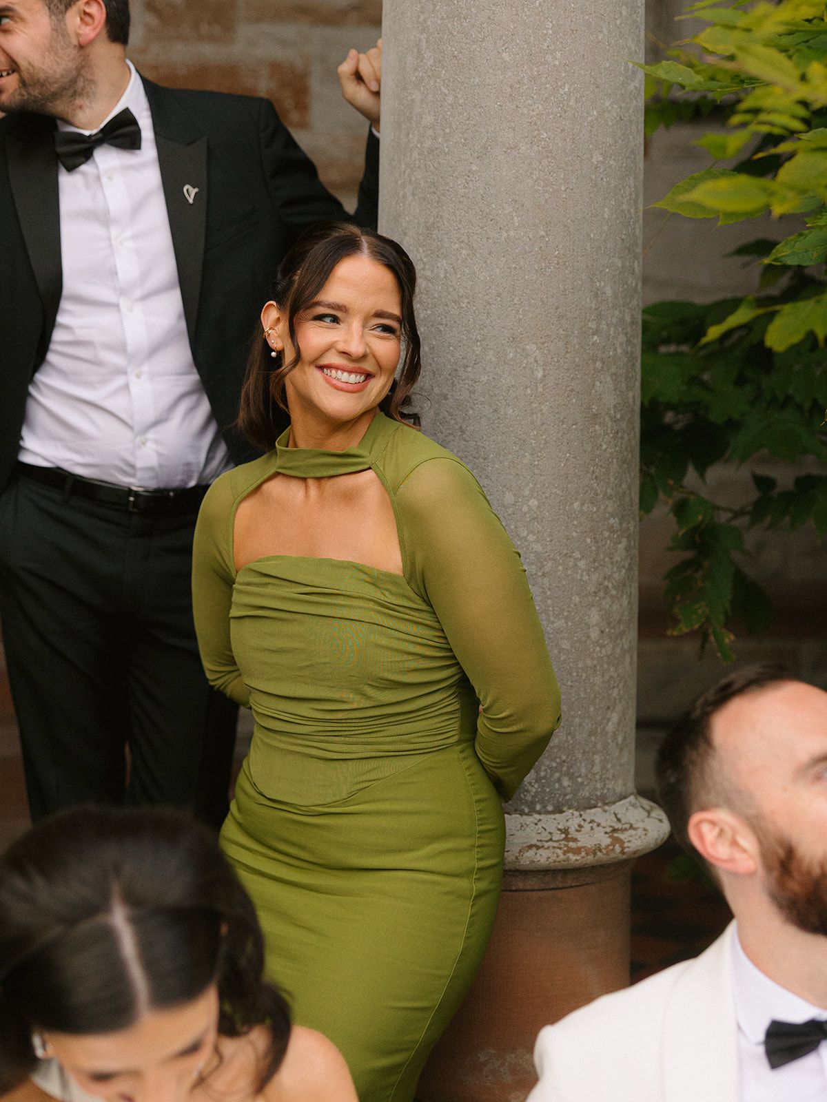 Woman in olive green dress smiling and posing at formal event with stone column in background.