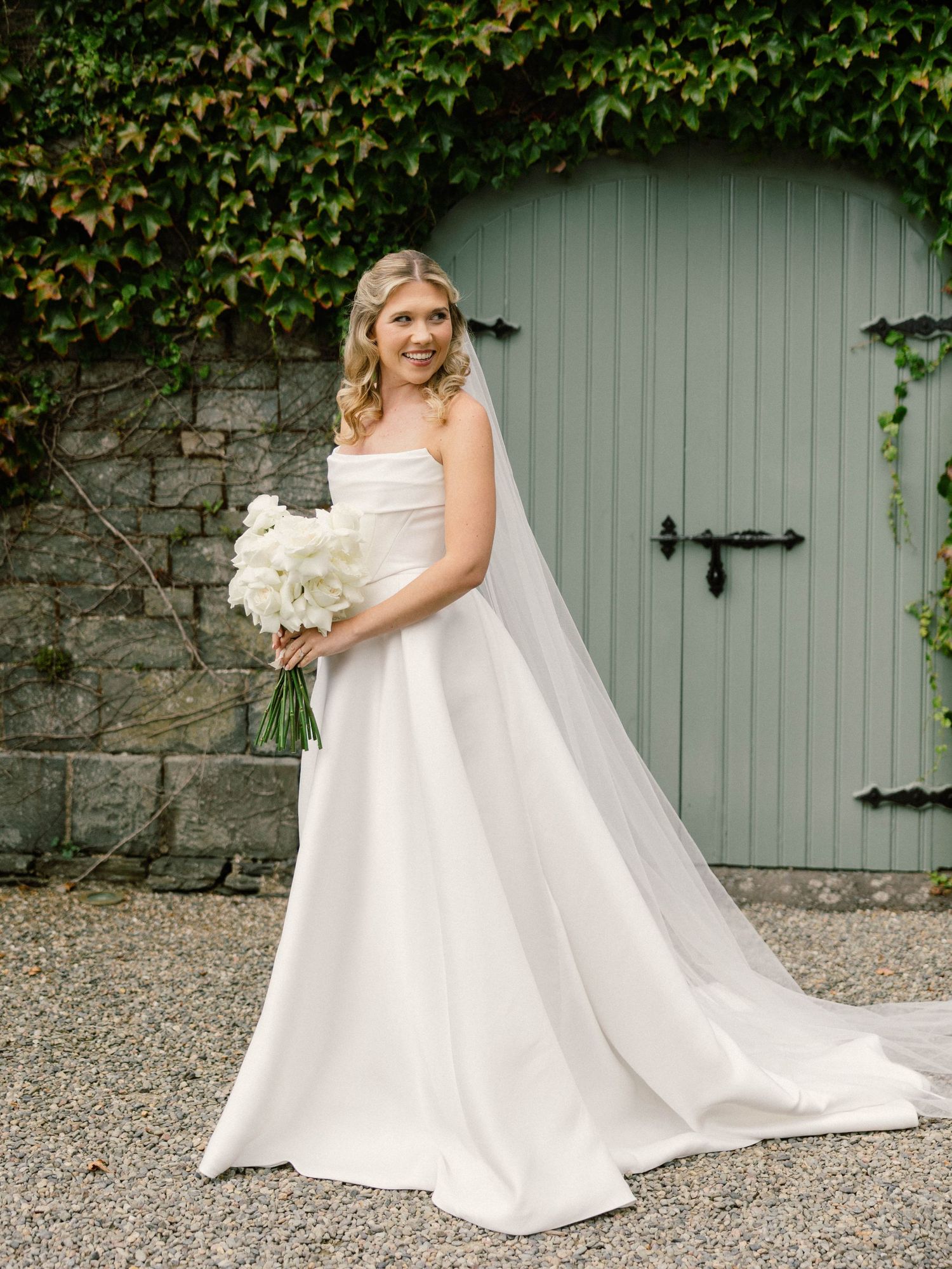 Elegant bride in flowing white gown against stone wall with ivy and mint green door.