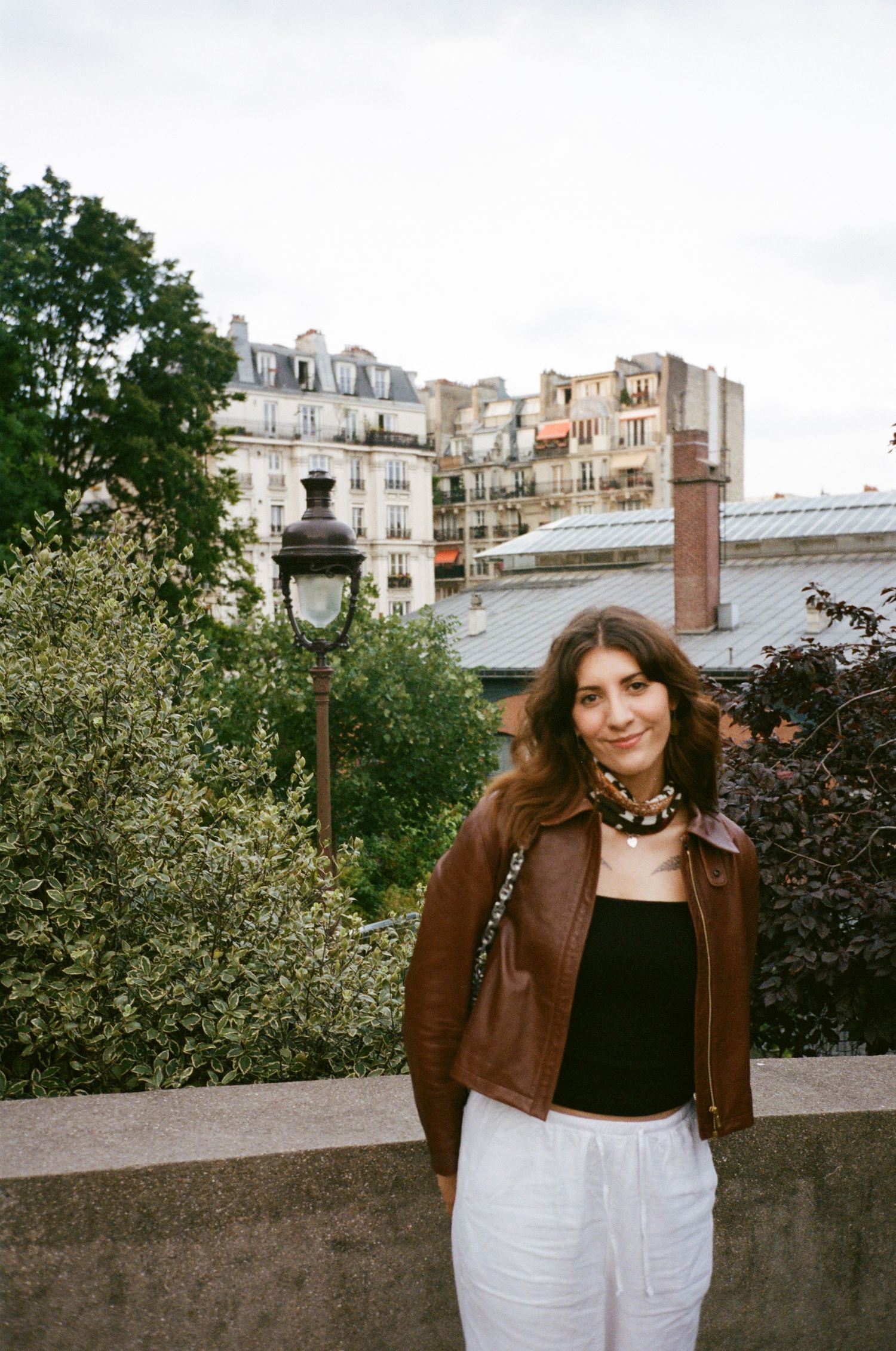 Person in brown leather jacket and white pants posing against urban Parisian backdrop.