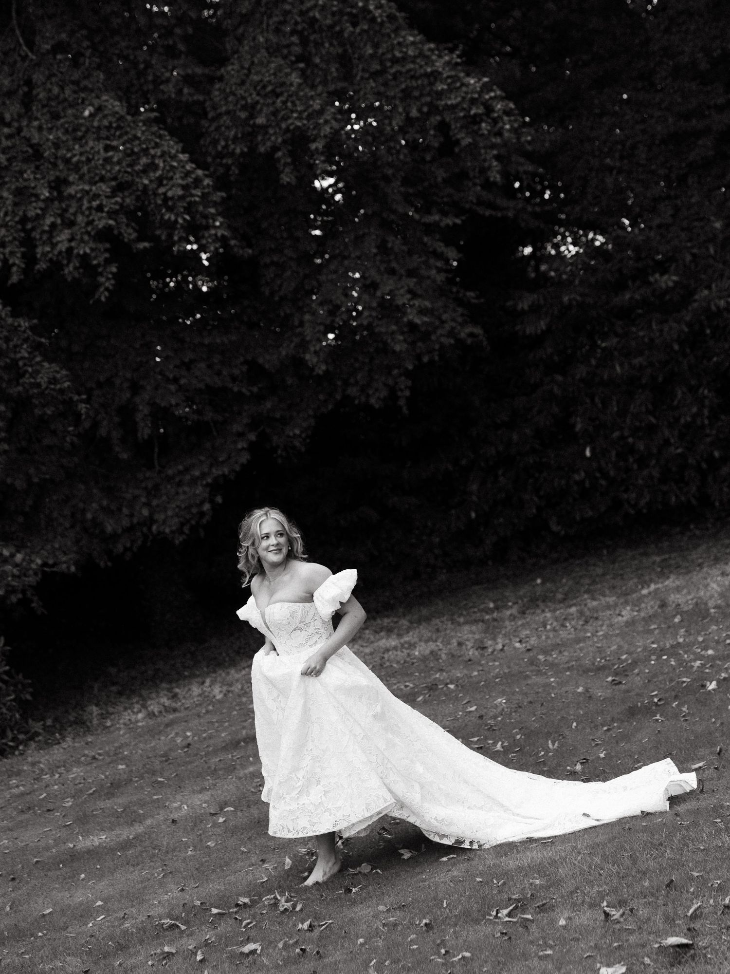 A bride in a flowing white gown walks gracefully across a grassy lawn surrounded by lush trees in a black and white photo.