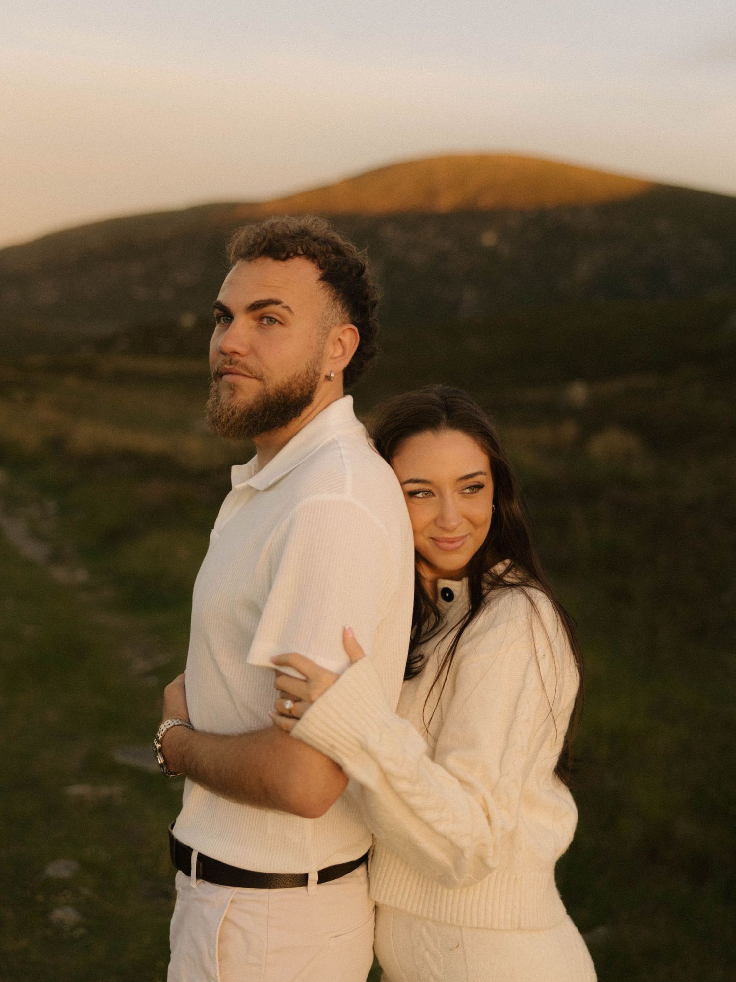Couple posing back-to-back at golden hour with rolling green hills and a mountain in the background.