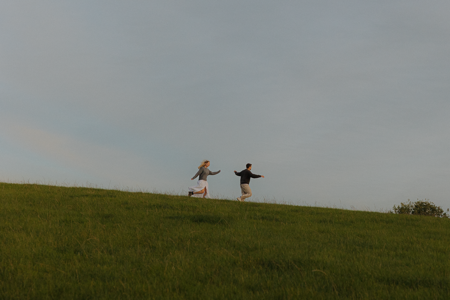 Couple holding hands running on a lush green hillside under a cloudy sky.