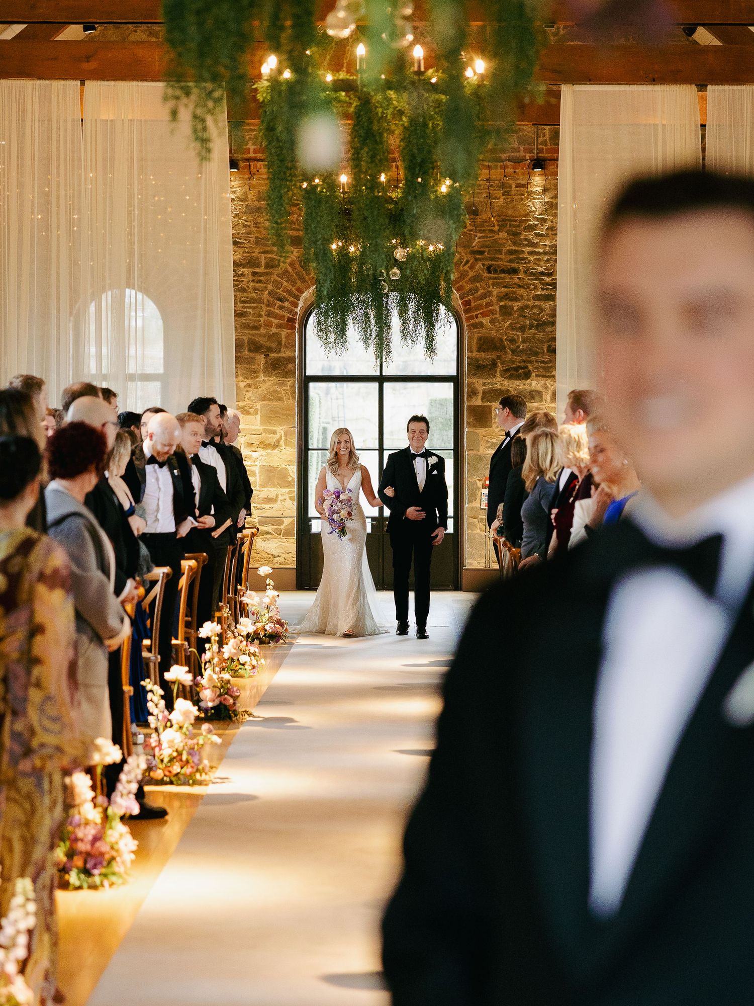 Elegant wedding ceremony aisle in rustic brick venue with string lights and floral arrangements lining the path.