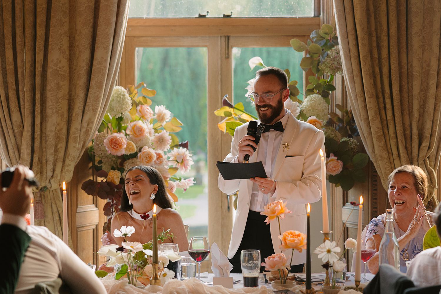 A wedding reception scene with a speaker giving a toast while guests sit at decorated tables with floral arrangements.