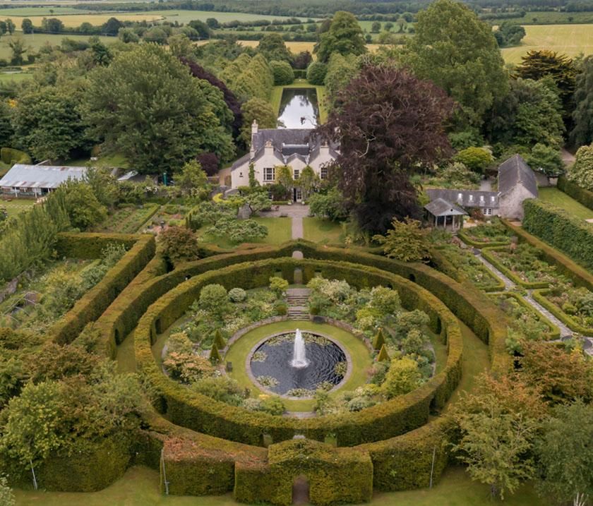 Aerial view of a circular garden maze with hedges and fountain centerpiece surrounded by lush greenery and countryside.