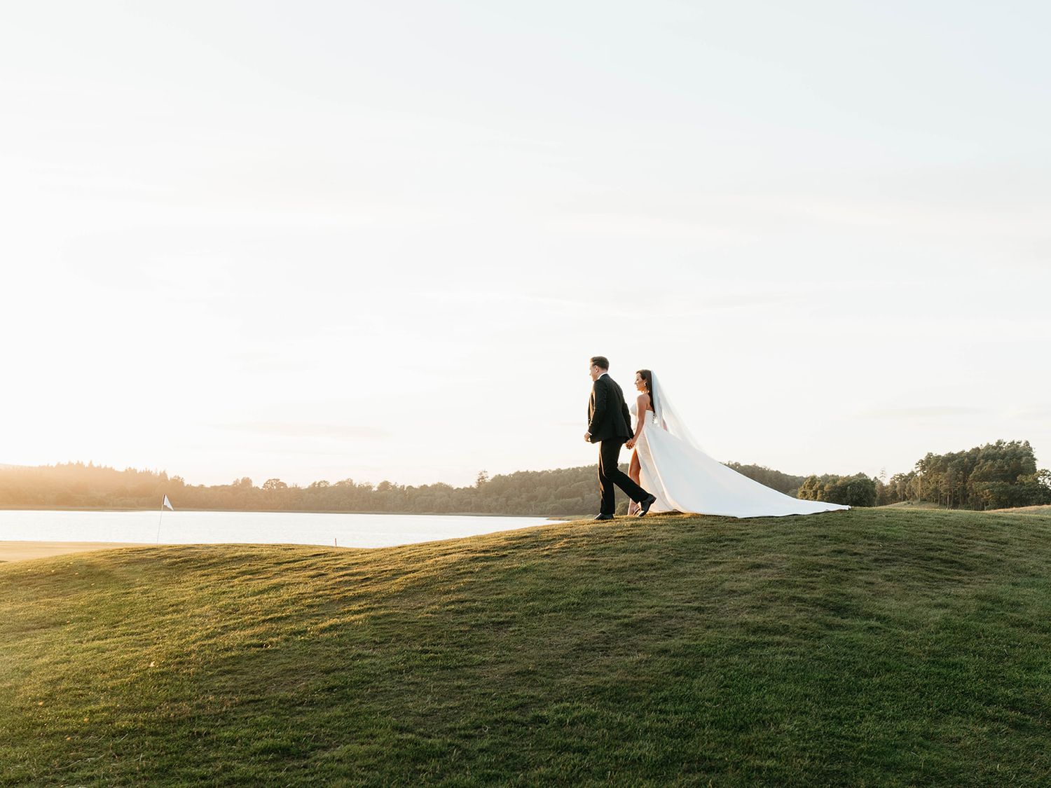 Silhouettes of people standing on grassy hill against sunset sky with lake in background.