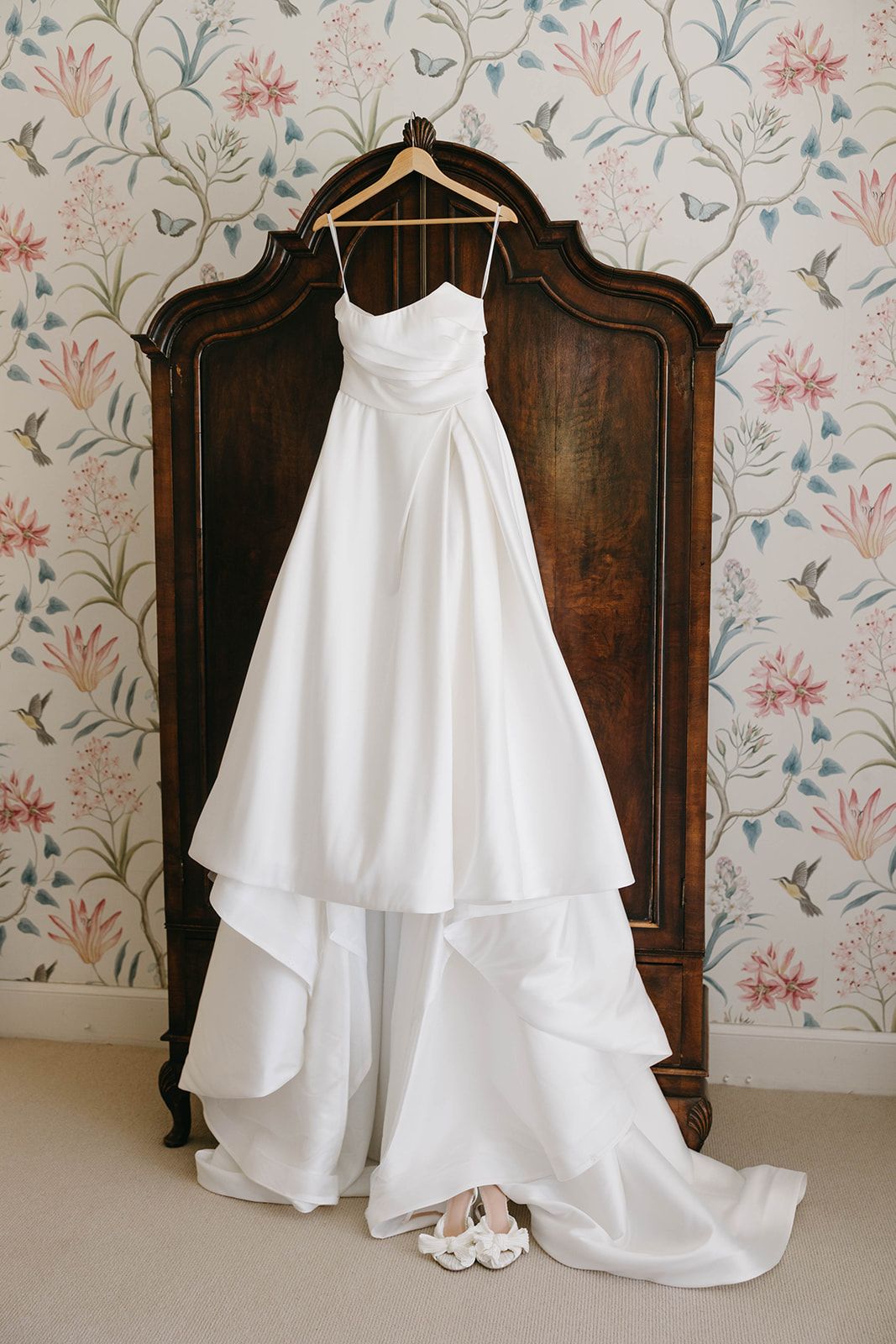 White wedding dress with layered ruffles displayed against vintage wooden furniture and floral wallpaper.