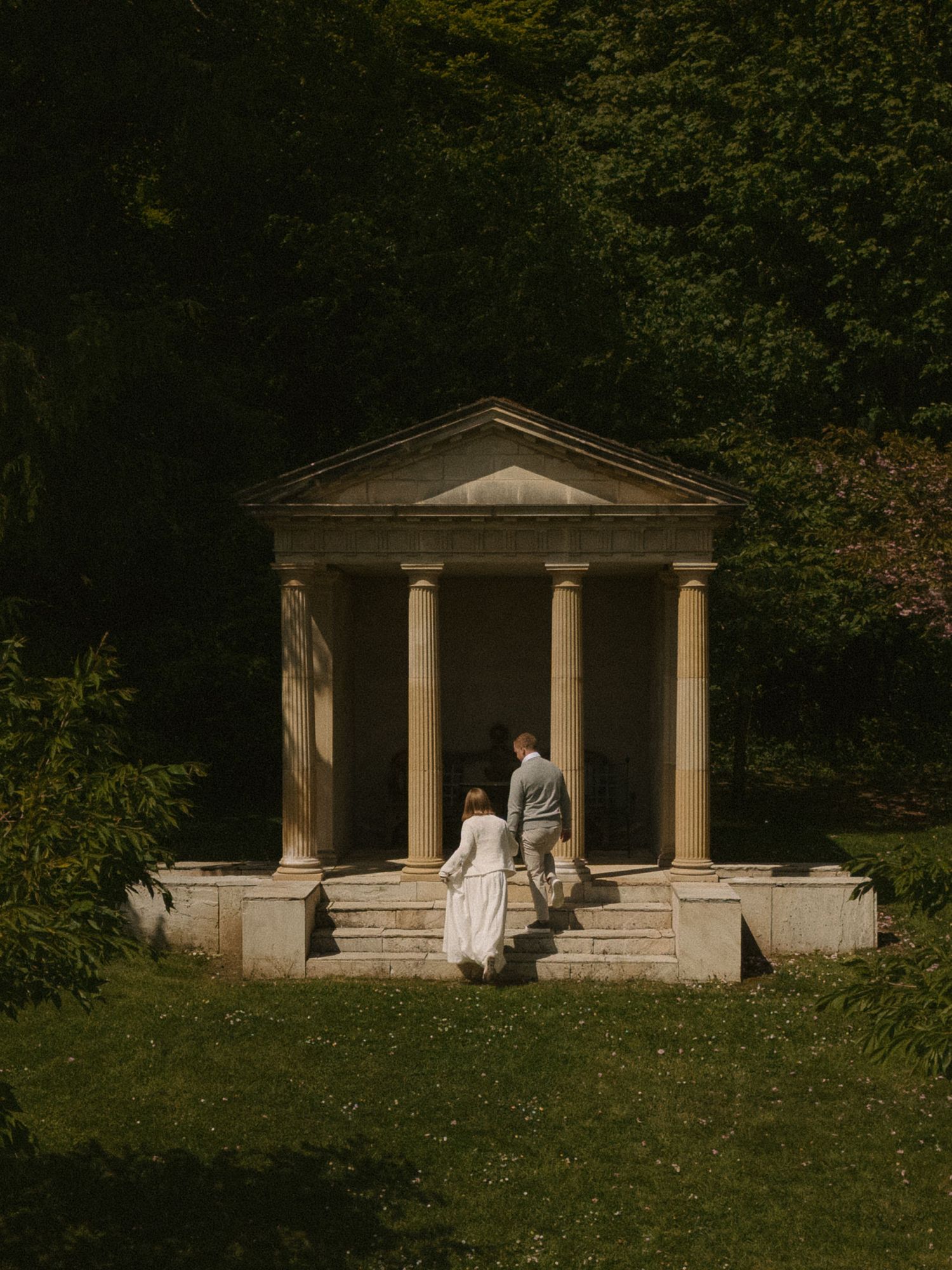 A couple walks hand-in-hand toward a neoclassical stone temple folly surrounded by lush green trees.