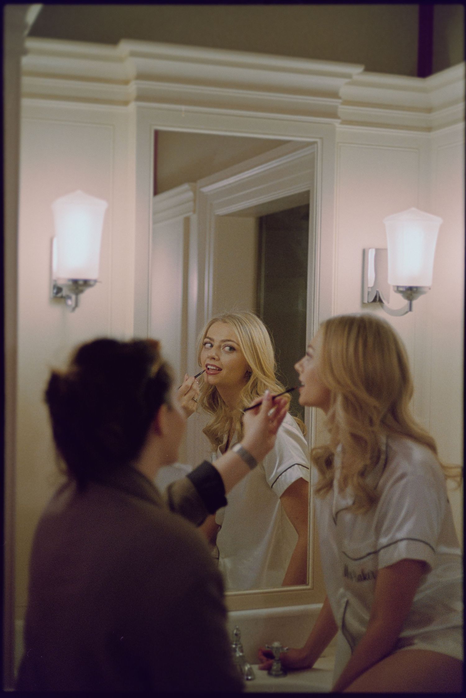A series of mirror reflections show someone applying makeup in a bathroom with white walls and sconce lighting.