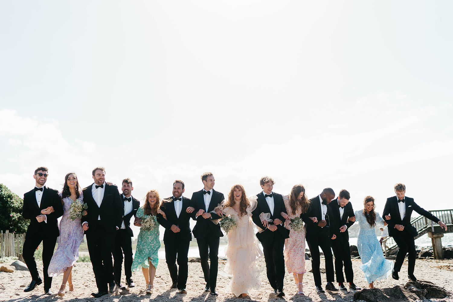 Wedding party members strike playful poses in a line on a sunny day outdoors.