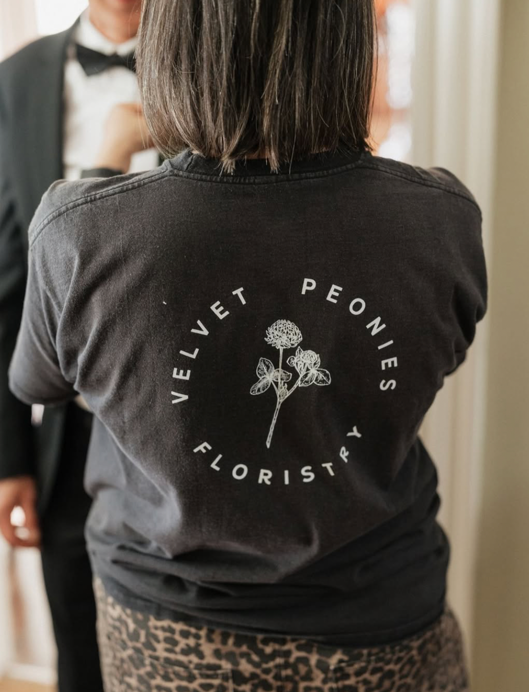 Person wearing black long sleeve shirt with floral design and text 'Vei-Lon Florists' printed on back.