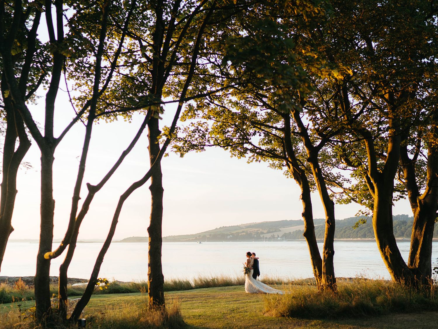 Silhouetted figures stand between trees at sunset overlooking a tranquil lake or ocean view.