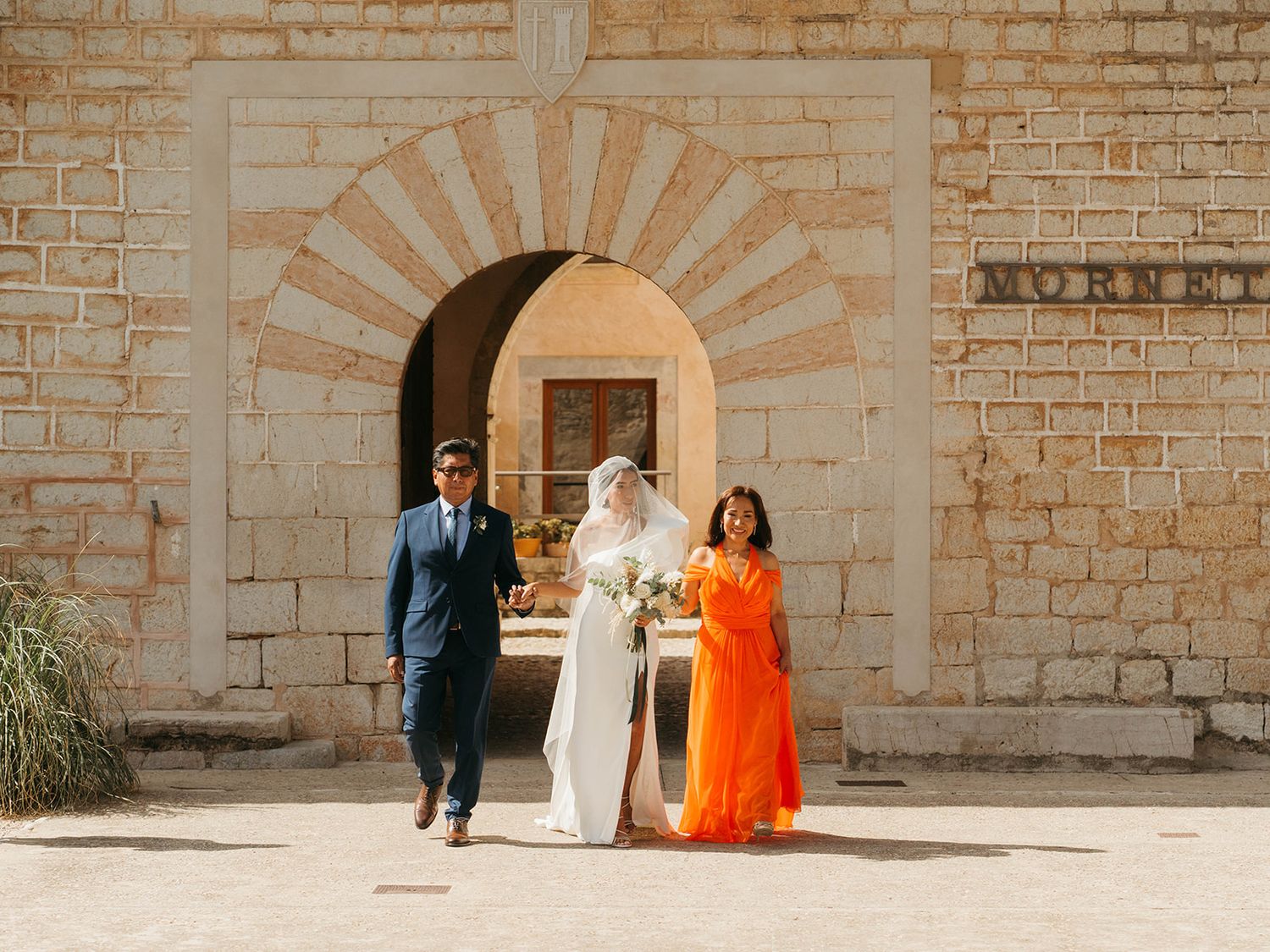 A wedding party walks through a stone archway while taking photos at a historic building.