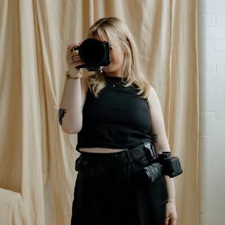 A blonde female photographer in black attire holds a camera in front of a beige curtain backdrop.