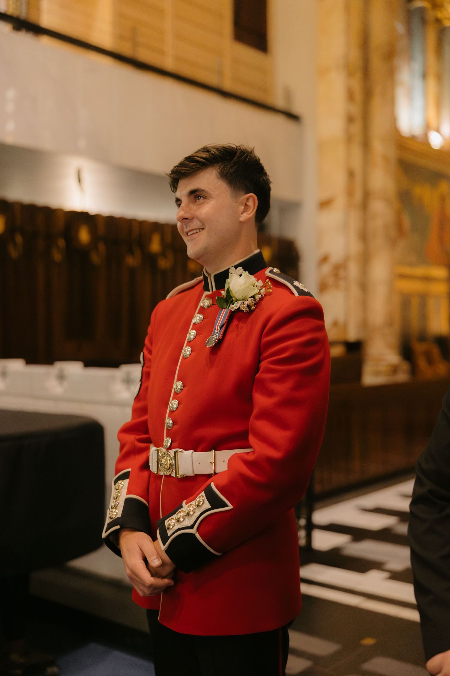 A person wearing a formal red military dress uniform sits in an elegant indoor setting.