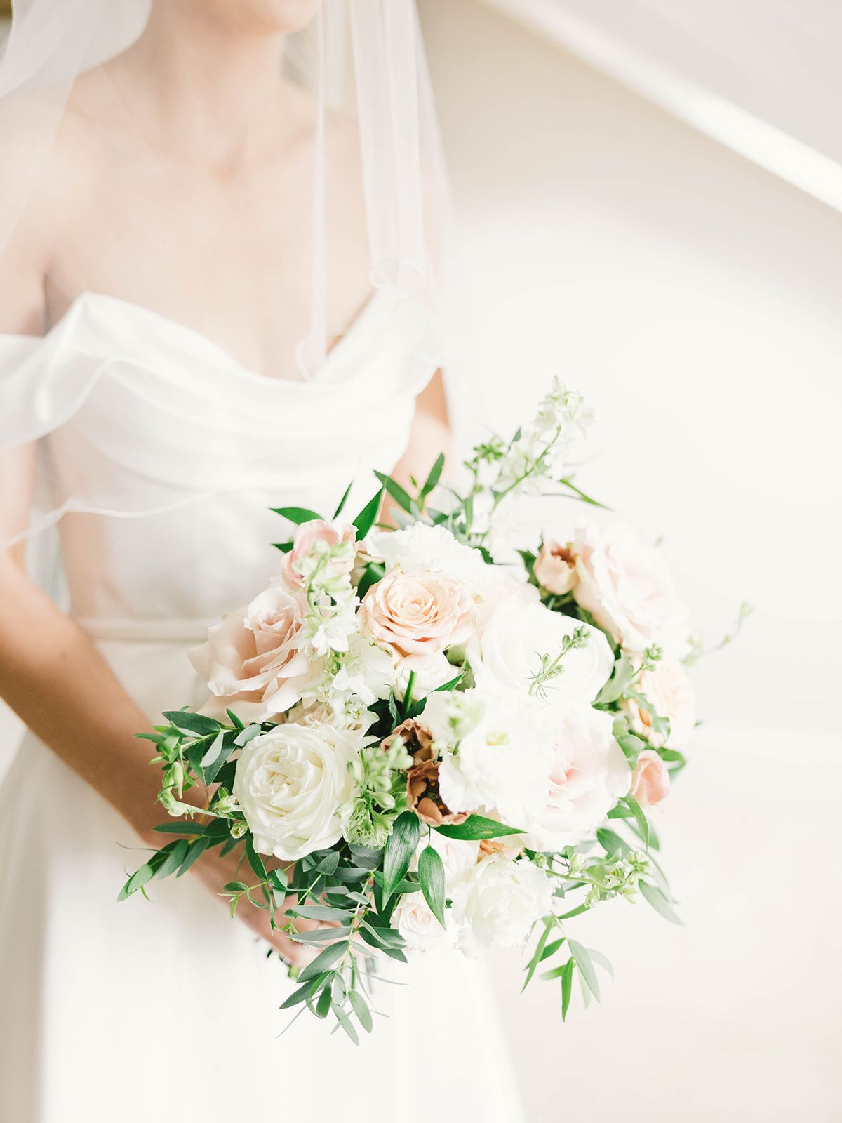An elegant white bridal bouquet features blush roses, white peonies and delicate greenery against a bright background.