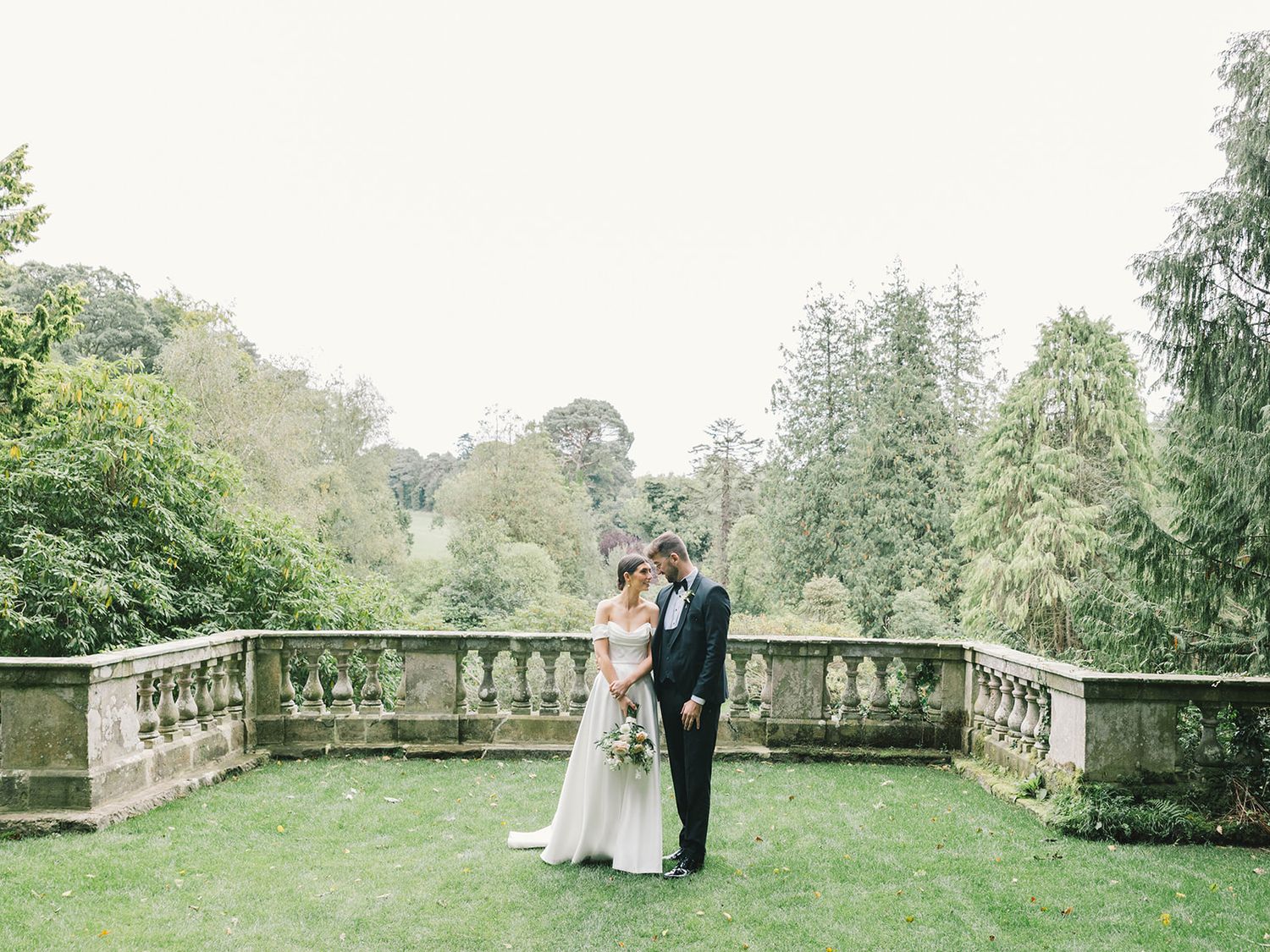 A couple shares a romantic moment on a stone terrace surrounded by lush green gardens and trees.