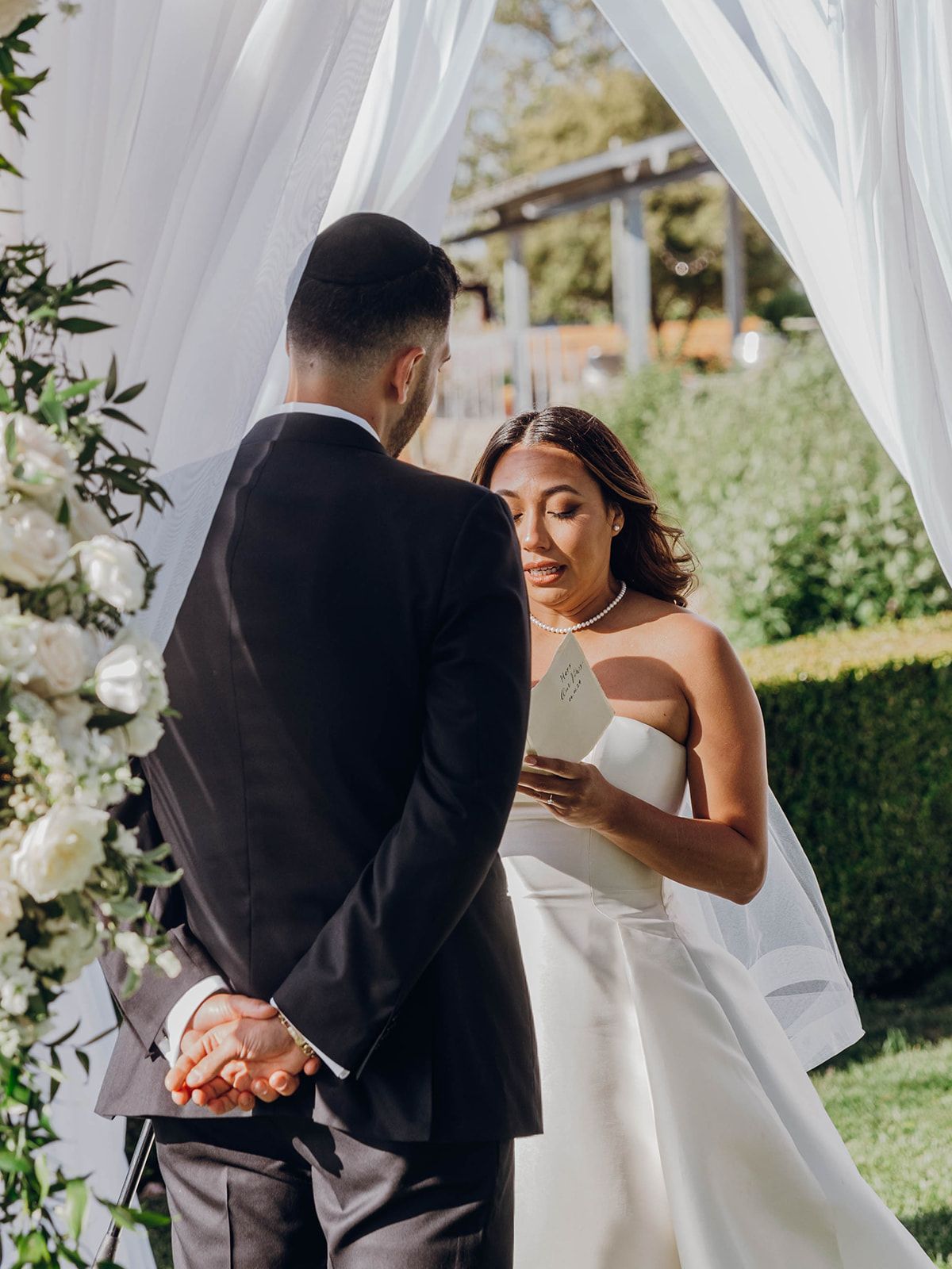 Emotional moment between couple at outdoor garden wedding ceremony with white flowing curtains and floral decorations.