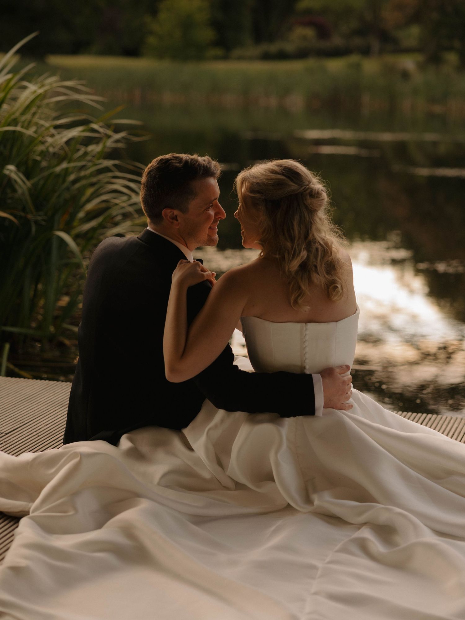 Romantic sunset photos of newlyweds sitting by a lake with flowing white wedding dress.